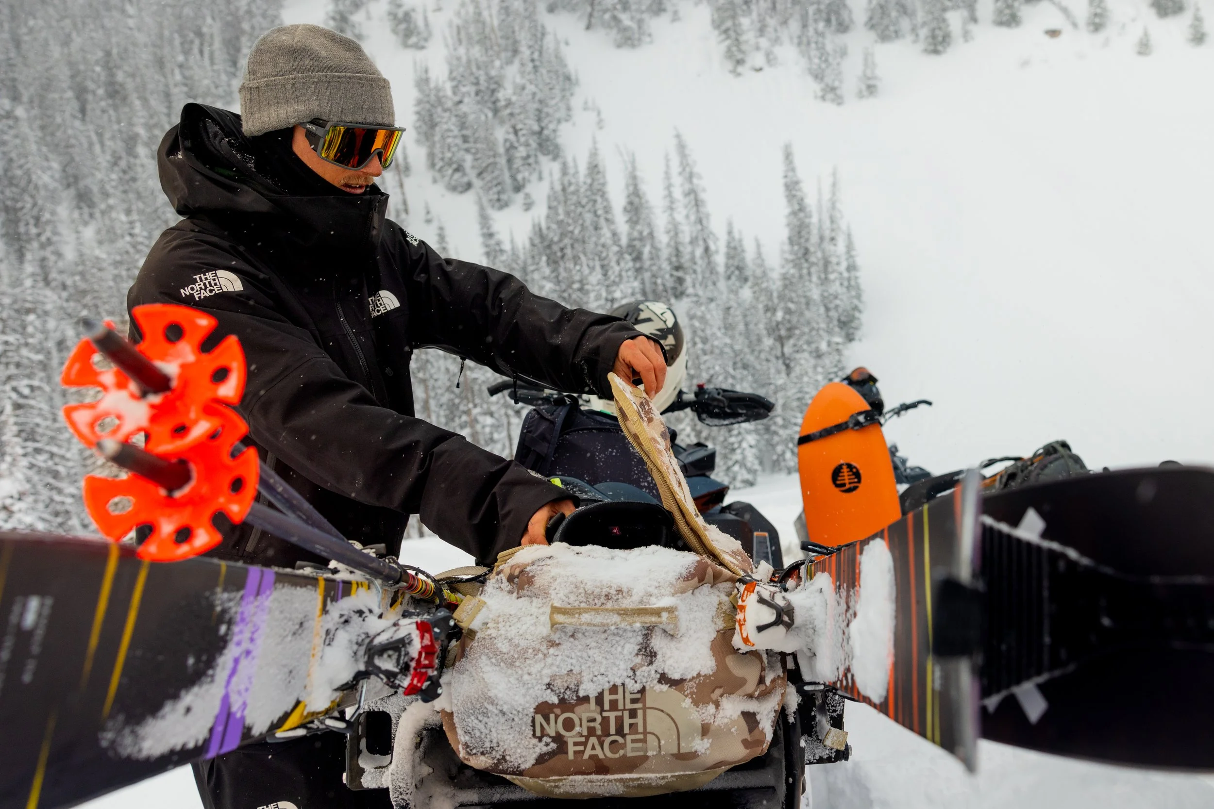 Man dressed in black winter jacket, gray beanie, and ski goggles, sitting on a snowmobile in snowy forested mountain landscape, with orange ski pole in foreground and a North Face duffle on the back of a snowmobile.