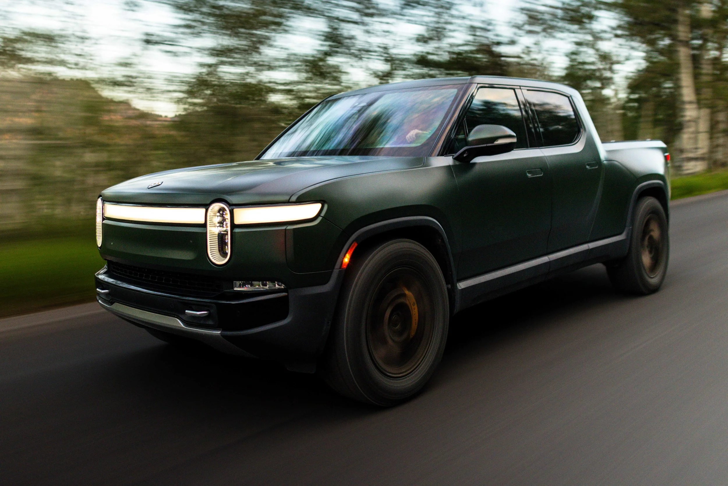 A matte green electric pickup truck driving on a road with a blurred background of trees, during daytime.