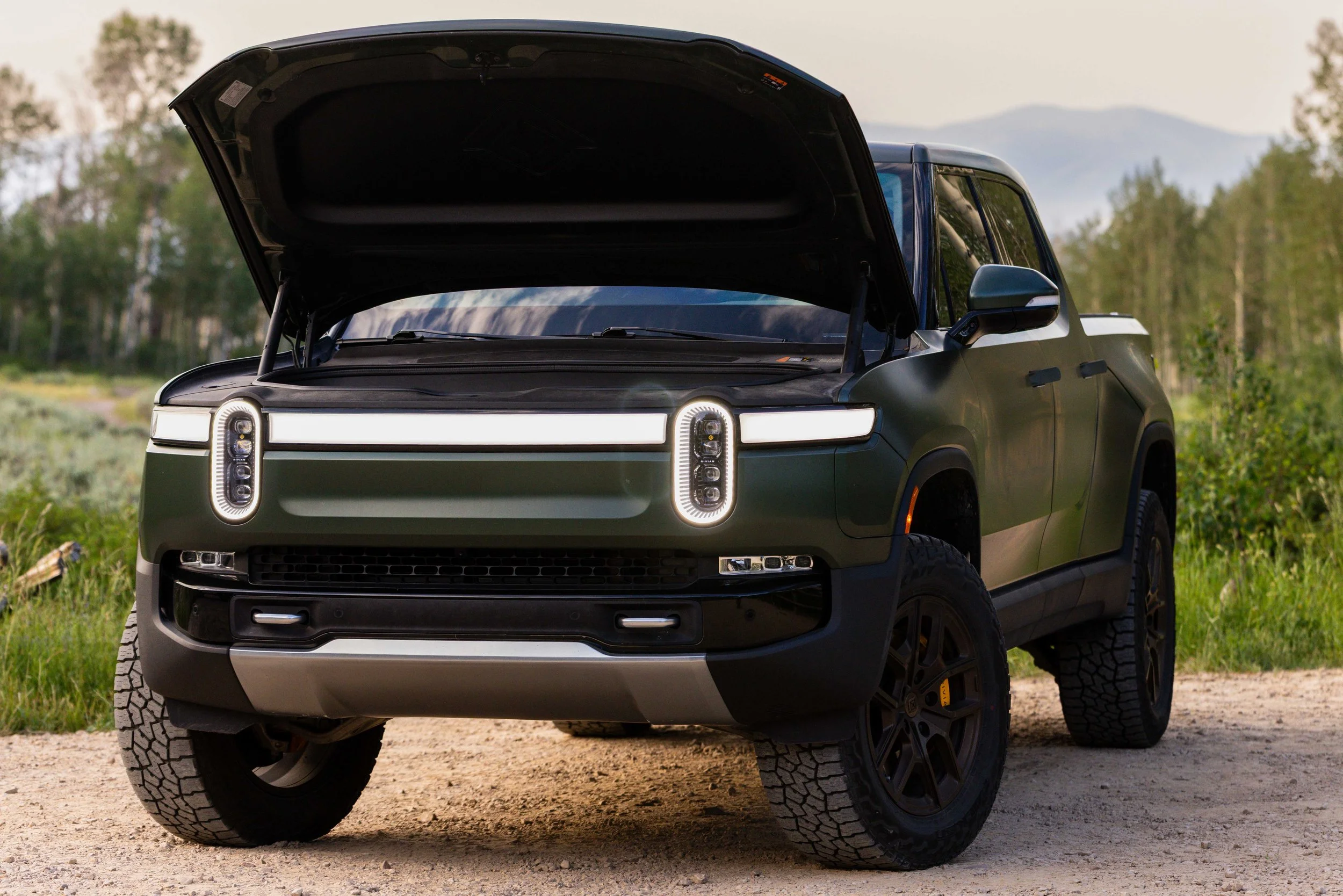 A green Rivian R1T truck with its hood open, parked on a dirt road in a natural landscape with trees and mountains in the background.