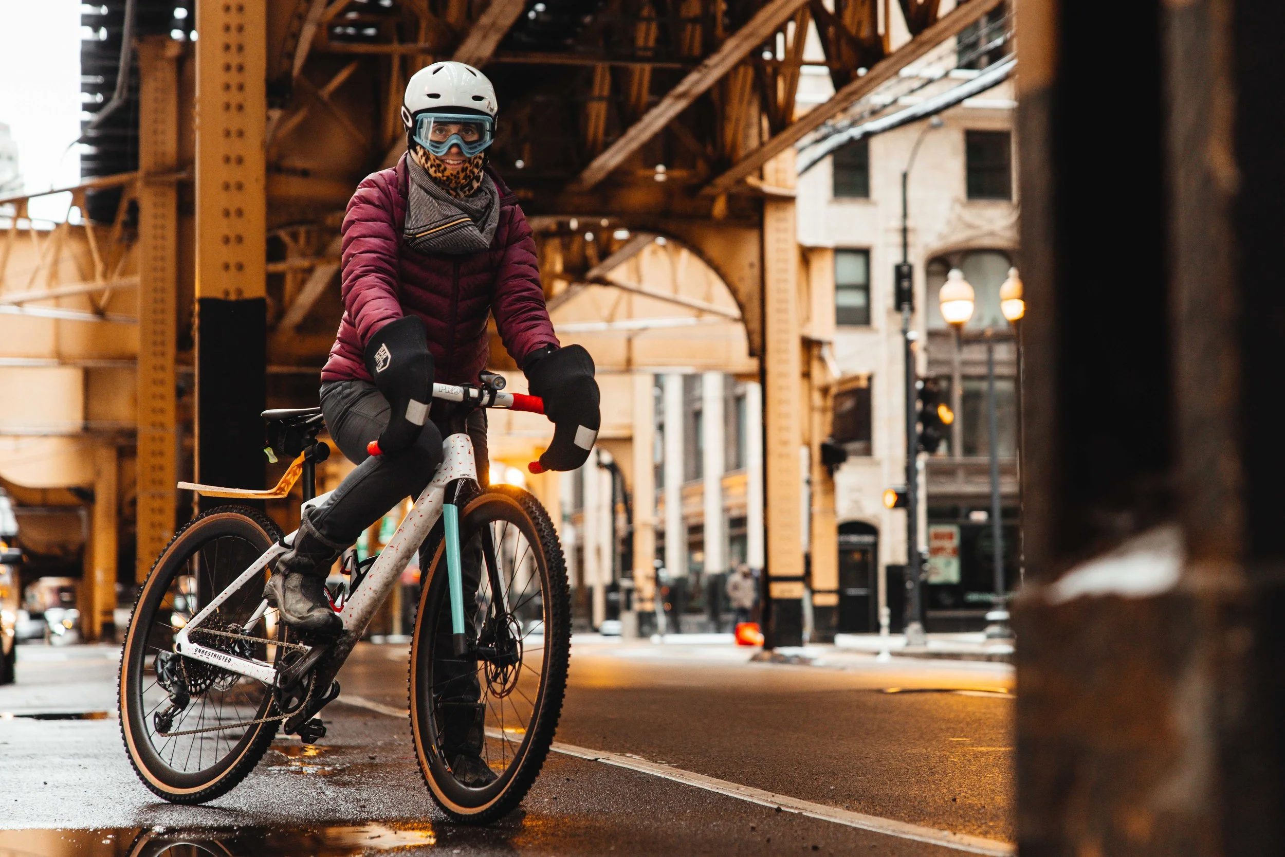 A person riding a BMC bicycle on a city street under a bridge, wearing a helmet, goggles, and a purple jacket.
