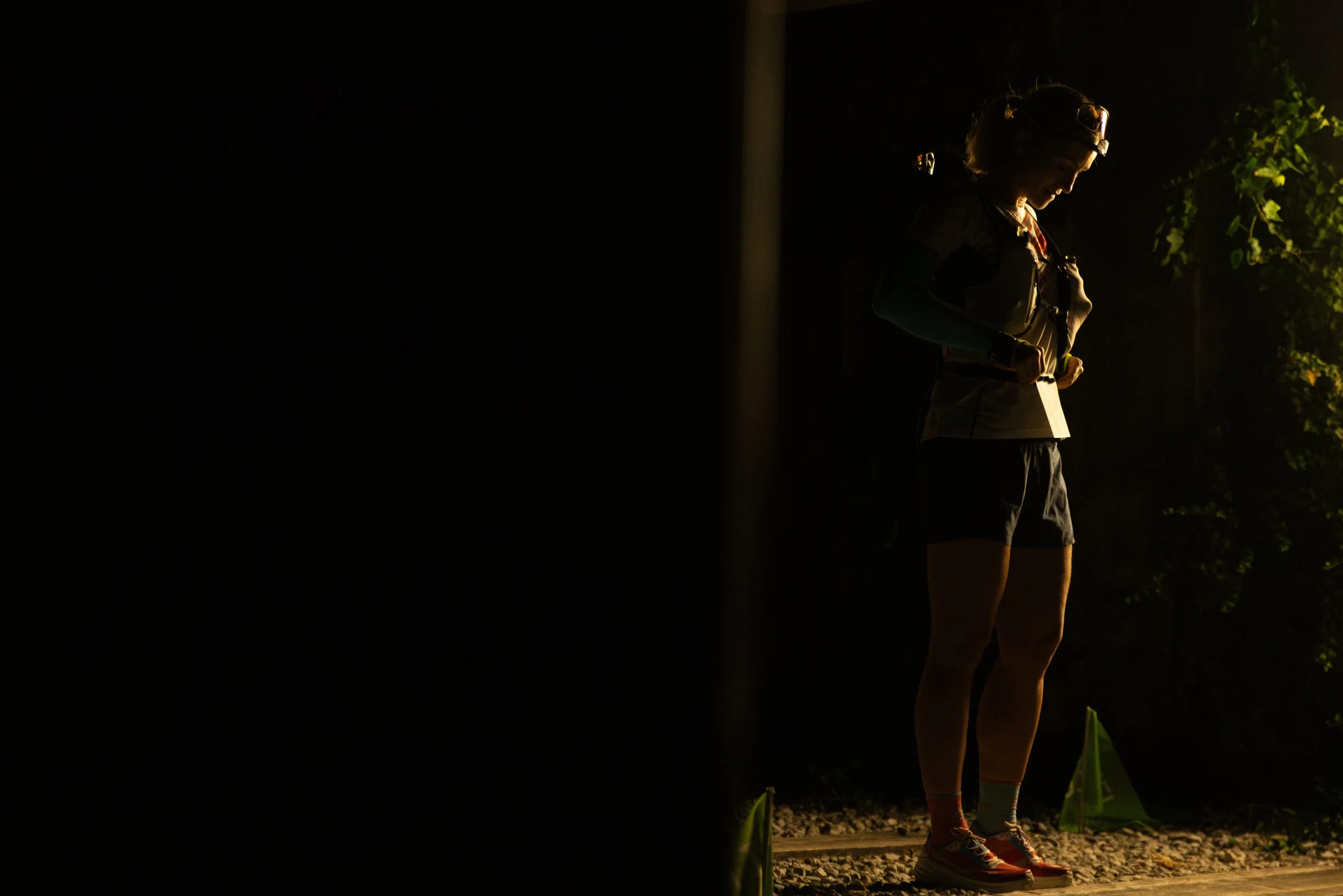 A woman standing outdoors at night, illuminated by a nearby light, wearing headphones and athletic clothing, possibly preparing for exercise.