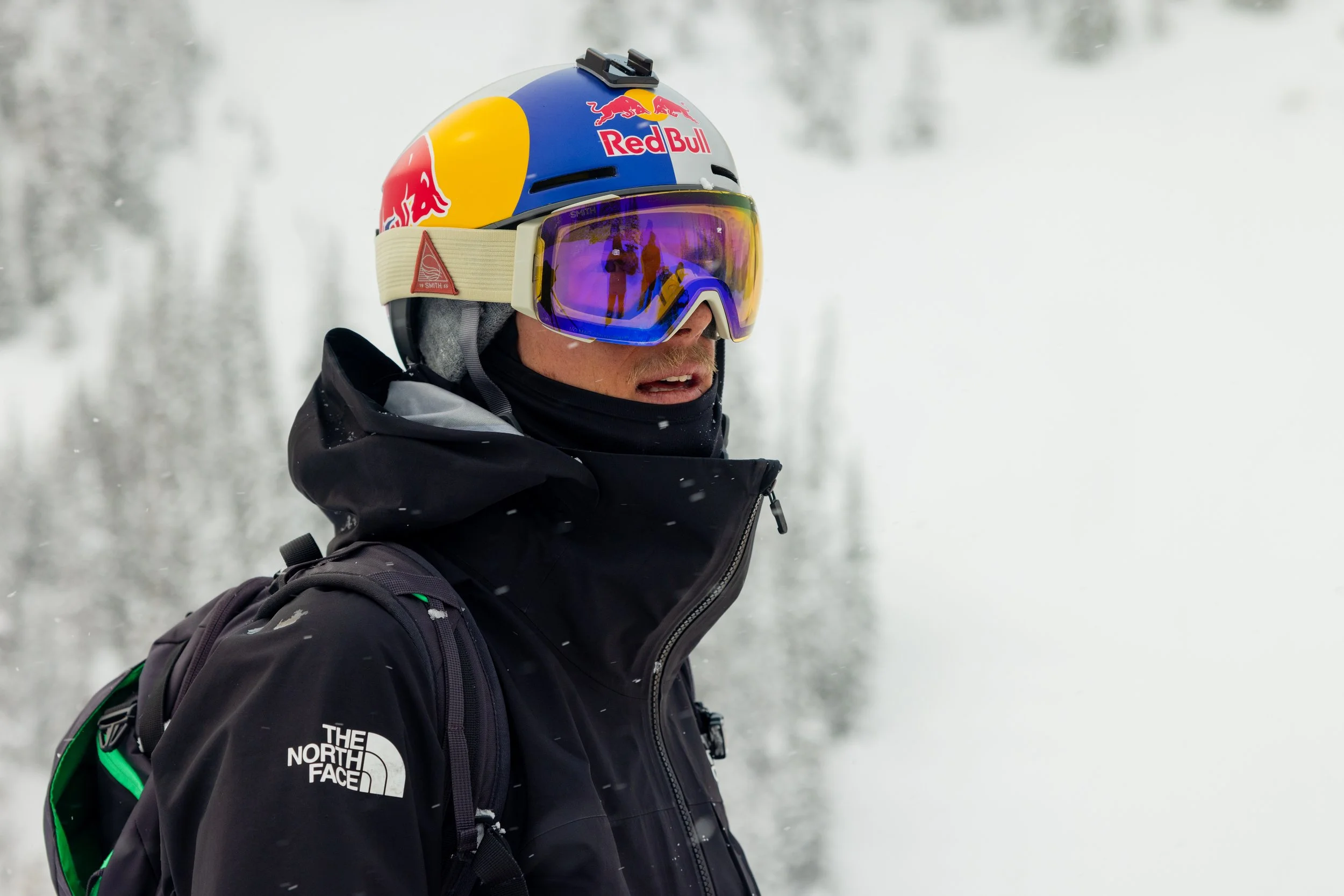 John Collinson wearing a ski helmet with a Red Bull logo, purple goggles, and a black North Face jacket, standing outdoors in a snowy environment.