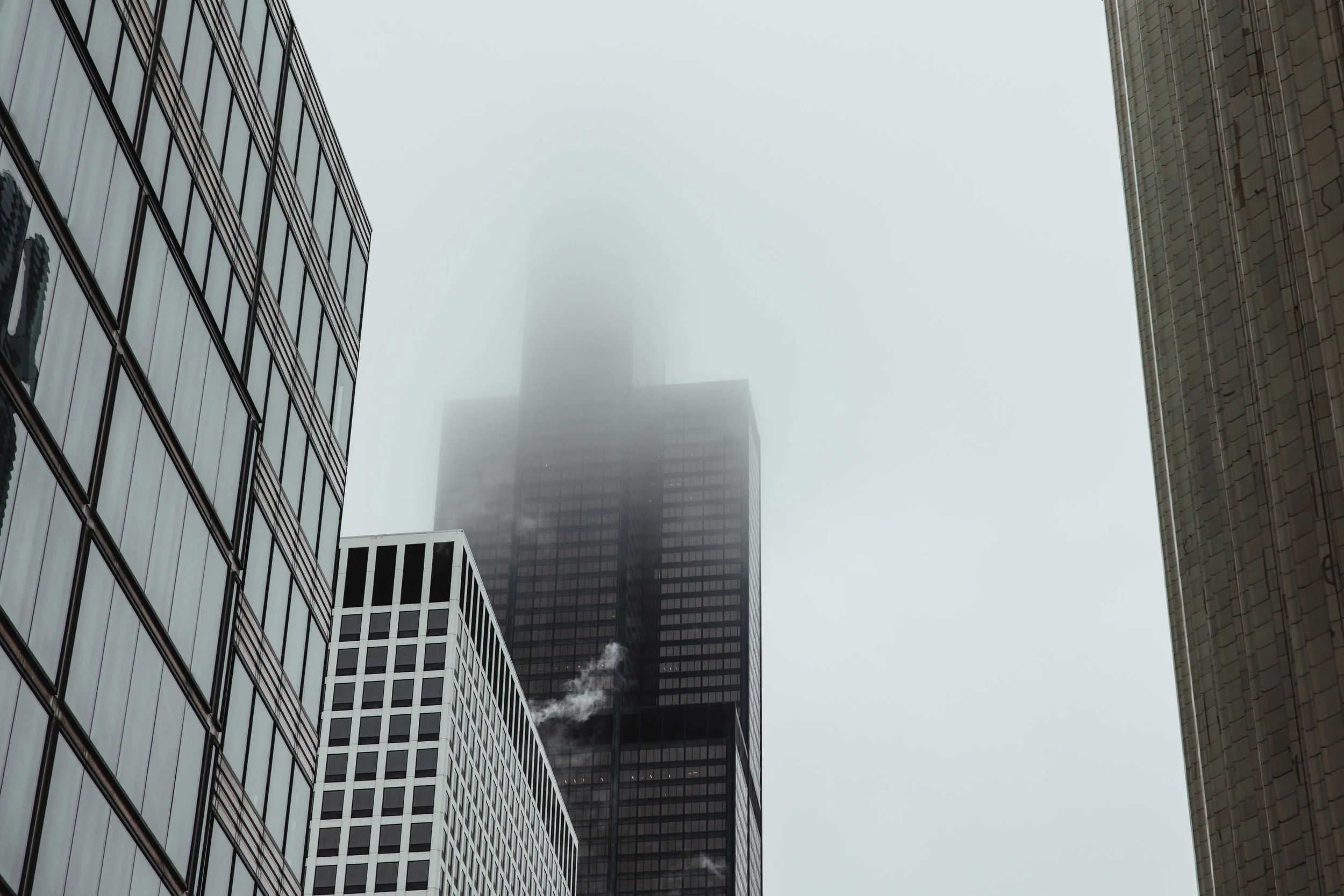 A Chicago city scene with tall buildings on a foggy day, partially obscuring a skyscraper in the distance emitting smoke or steam from its top.