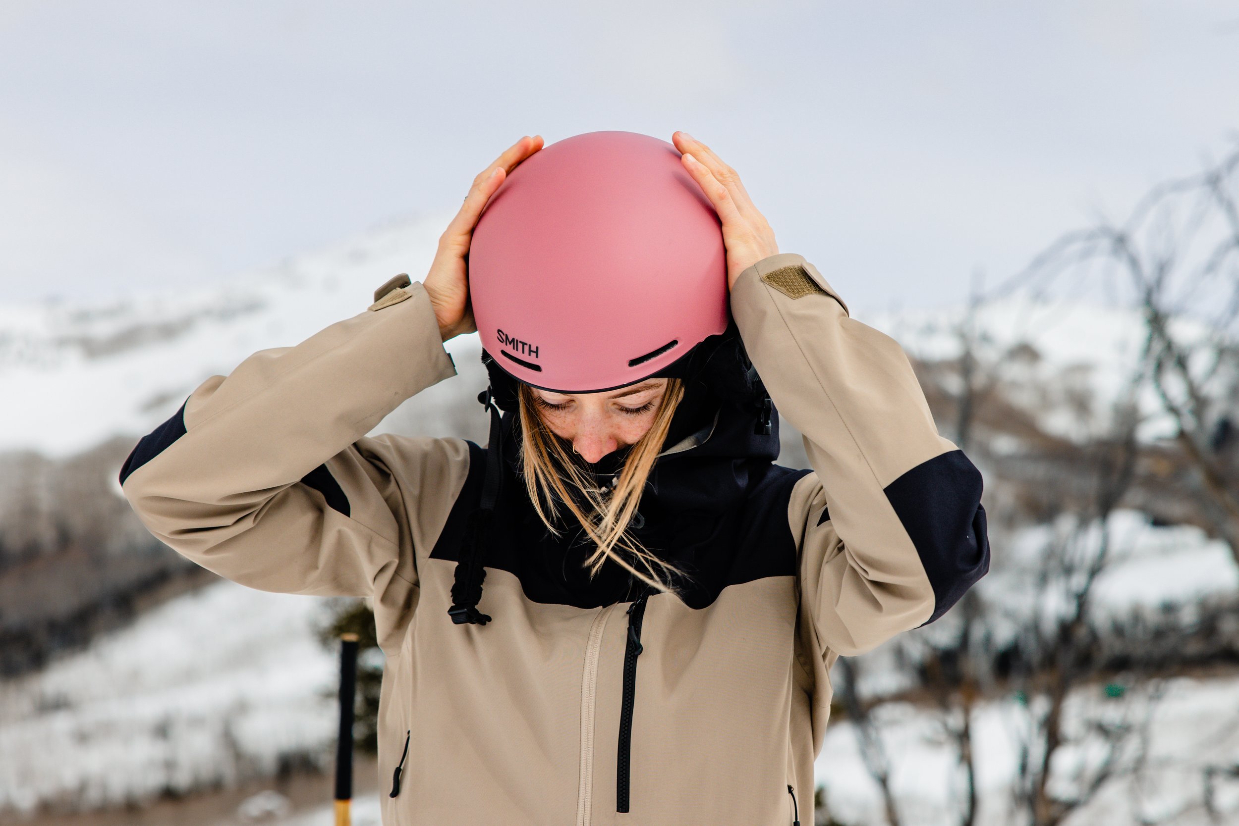A person in outdoor winter clothing holding a pink helmet on their head in a snowy outdoor setting.
