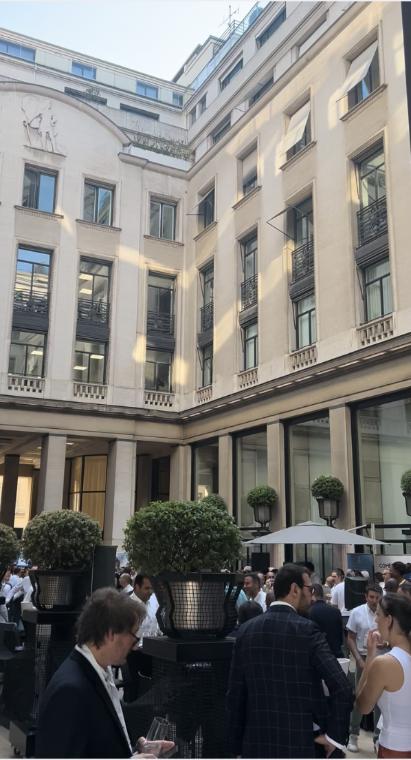 Outdoor courtyard of a historic building with people attending a social event, large potted plants, and an umbrella, with tall windows and architecture features.