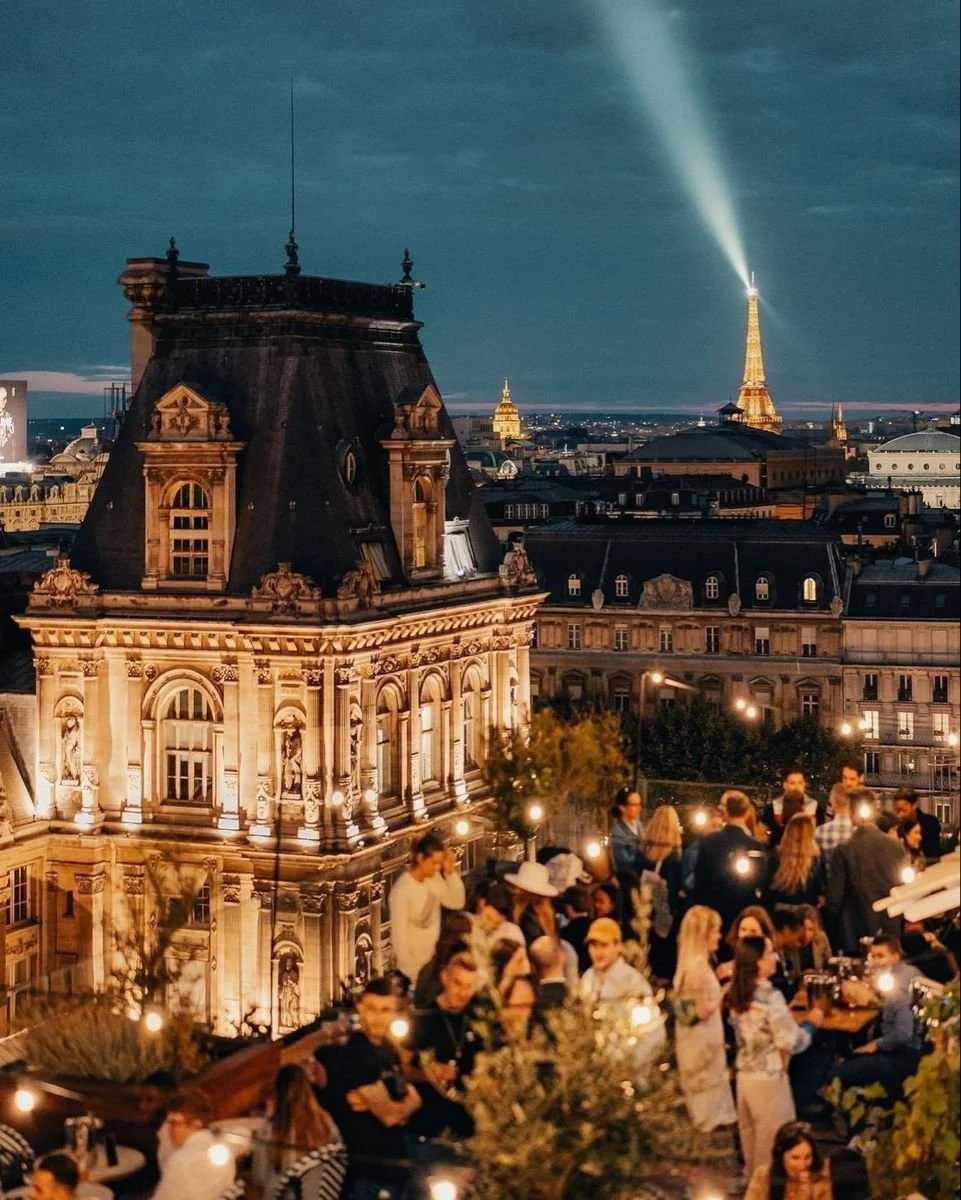 People dining at an outdoor rooftop restaurant in Paris at night, with illuminated historic buildings and the Eiffel Tower in the background.