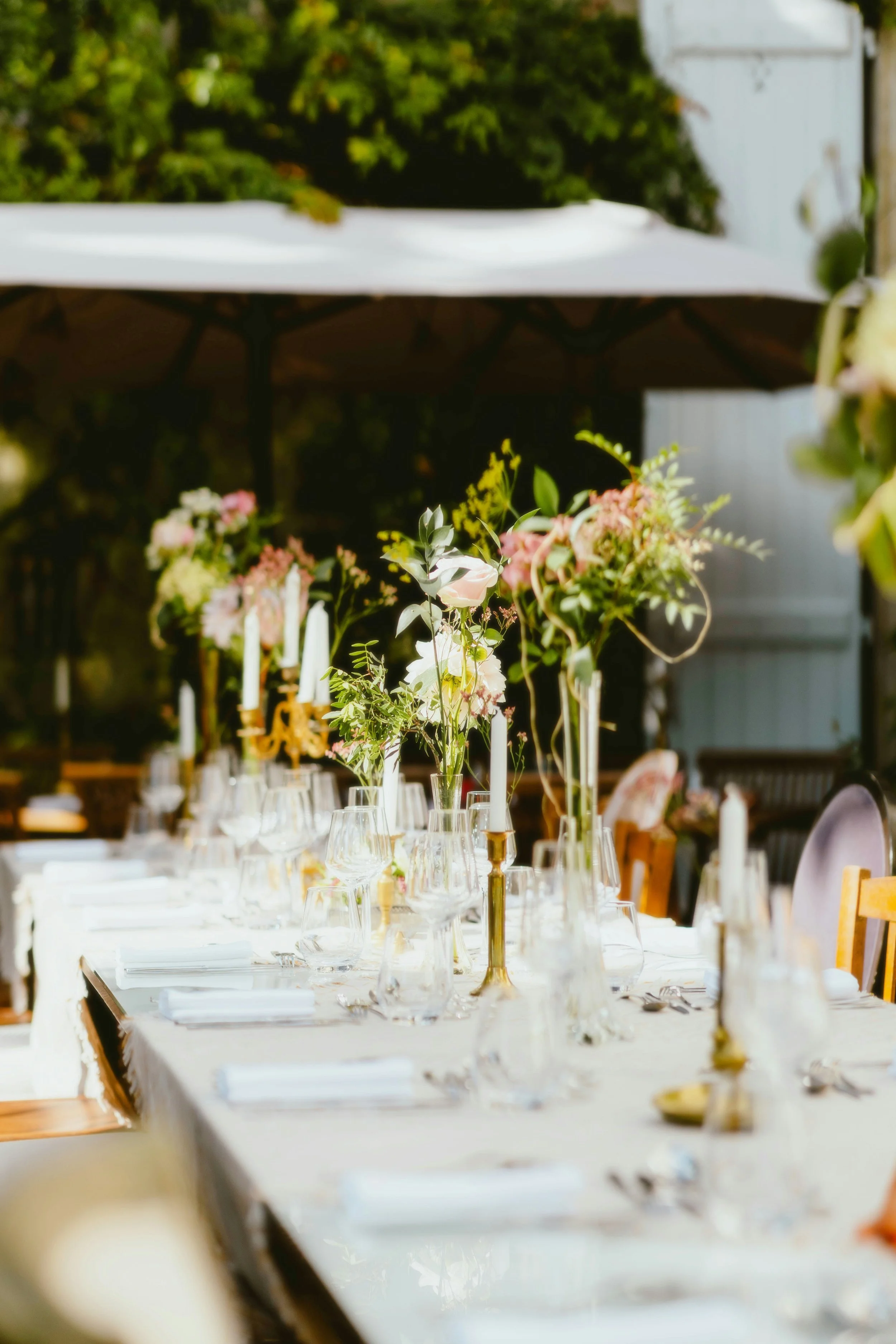 A long dining table set for a formal event outdoors, decorated with floral centerpieces and candles under a large white umbrella.