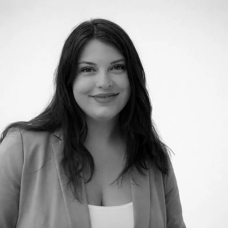 Black and white photo of a woman with long dark hair, smiling, wearing a blazer and a white top, standing against a plain background.