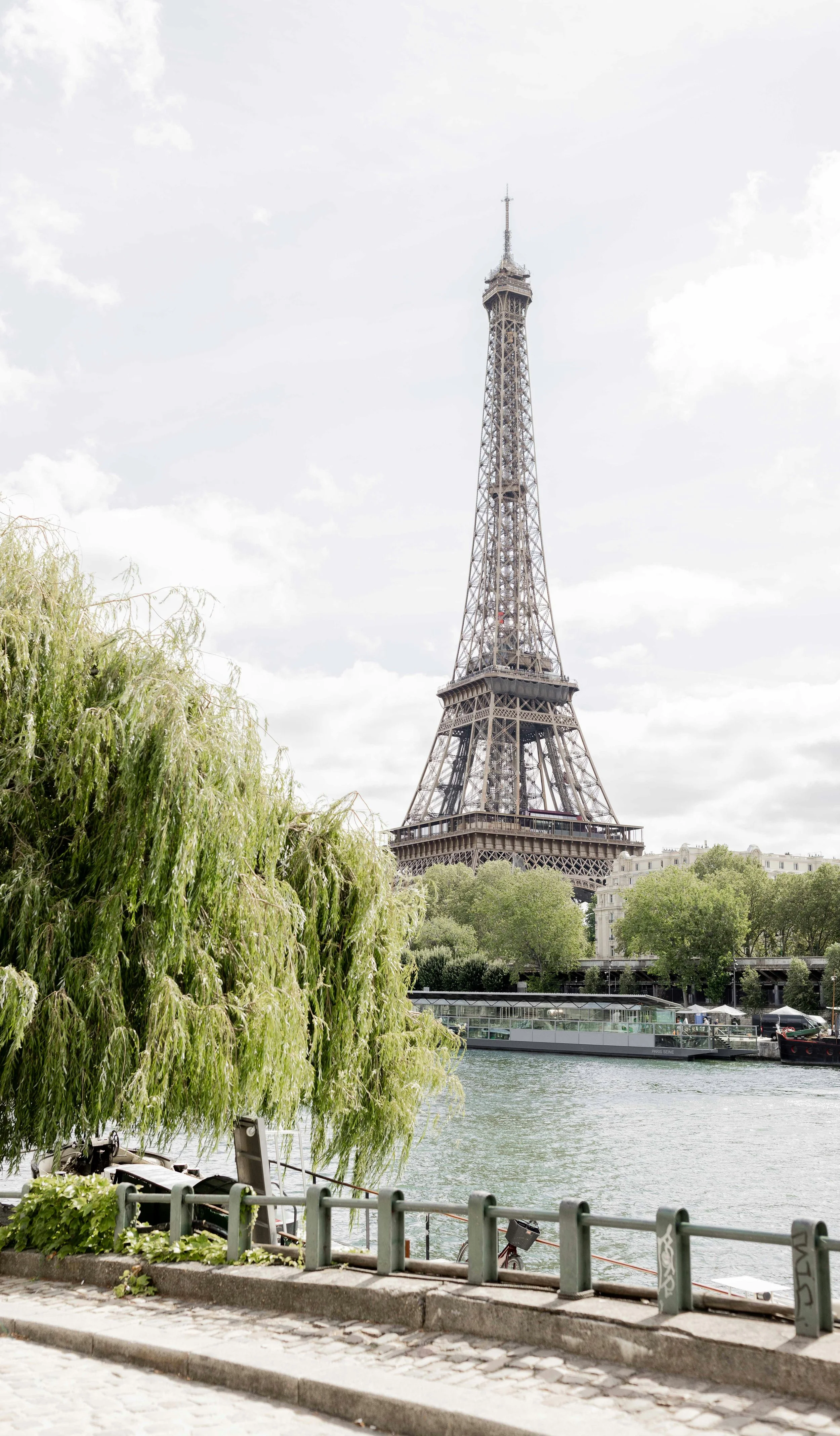 View of the Eiffel Tower seen from across the Seine River with greenery and a cobblestone walkway in the foreground.