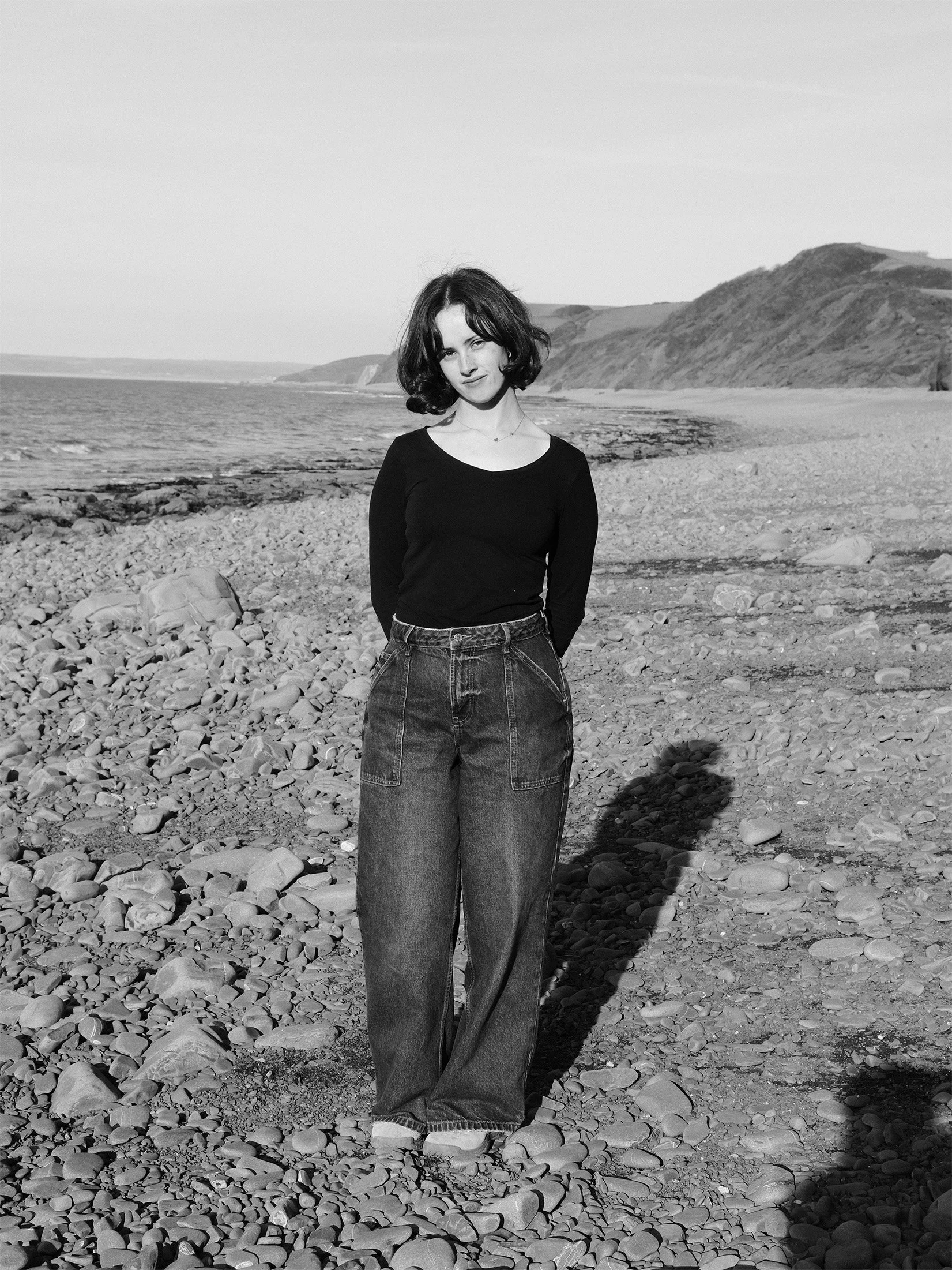 A young woman with short, wavy hair standing on a rocky beach with large stones, facing the camera with a slight smile, hillside and ocean in the background, in black and white.