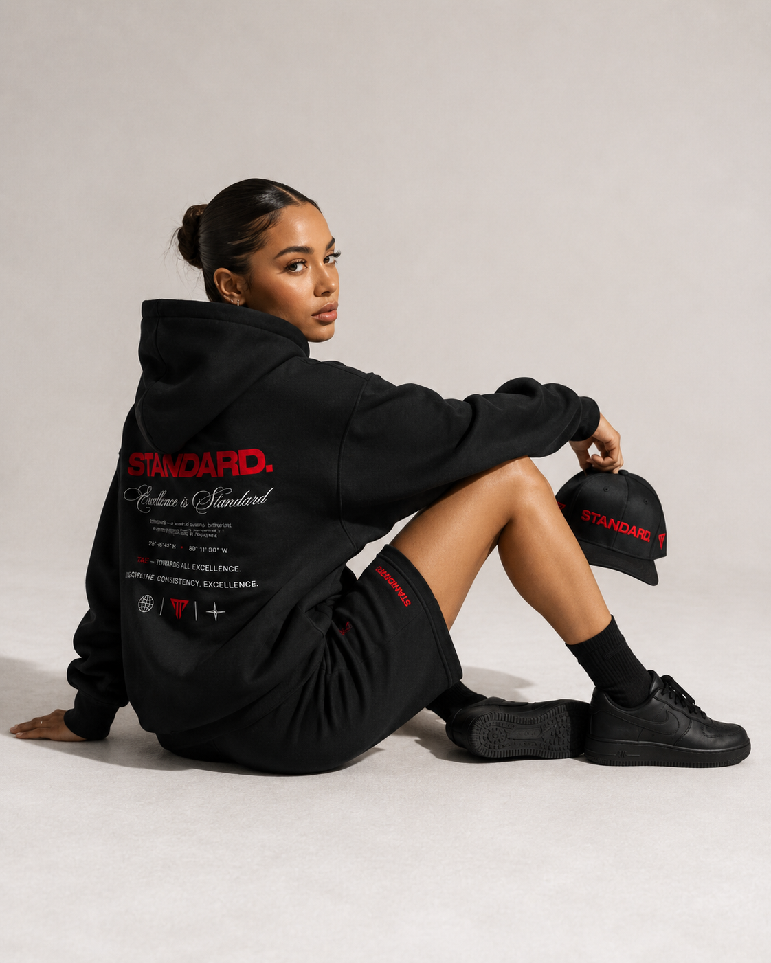 A young woman sitting on the floor in casual streetwear, wearing a black hoodie with red and white text, black shorts, black sneakers, and holding a black cap with red text