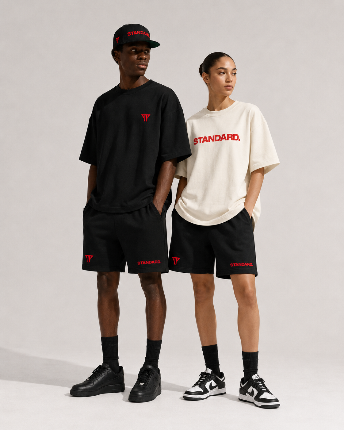 Two young adults, a man and a woman, standing against a plain background, wearing casual athletic outfits with black shorts, black and white sneakers, black socks, and T-shirts with the word 'STANDARD.' printed in red, and the man also wearing a black cap.