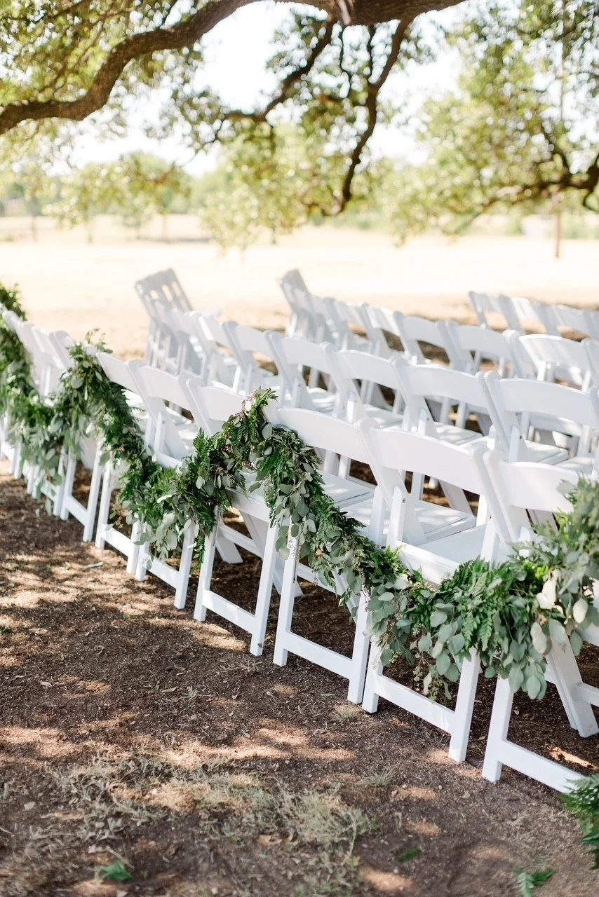 White chairs decorated with green foliage garlands arranged outdoors under a tree for a wedding ceremony.