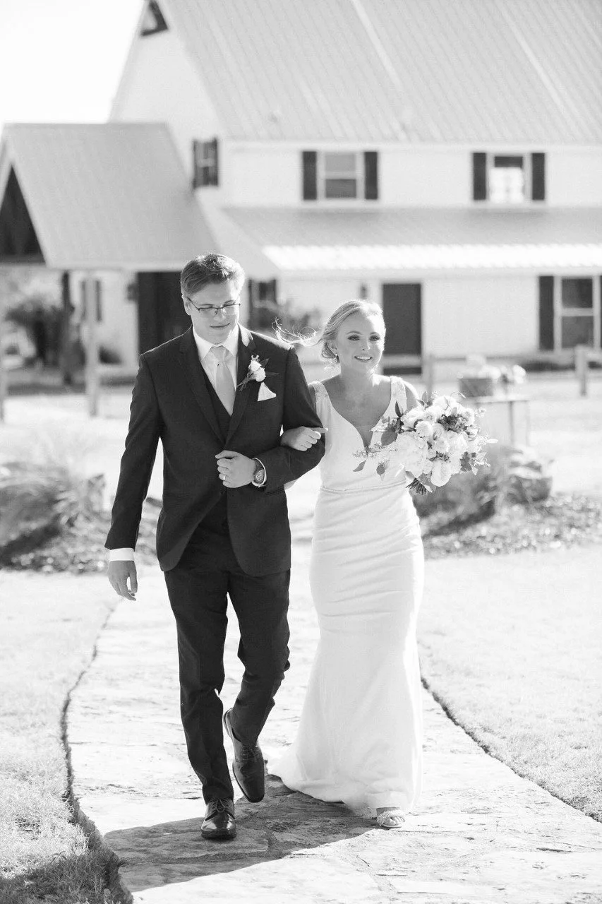 A black-and-white photo of a bride and groom walking arm-in-arm outdoors, with the bride smiling and holding a bouquet of flowers, and the groom looking down. They are in front of a house with distinct rooflines.