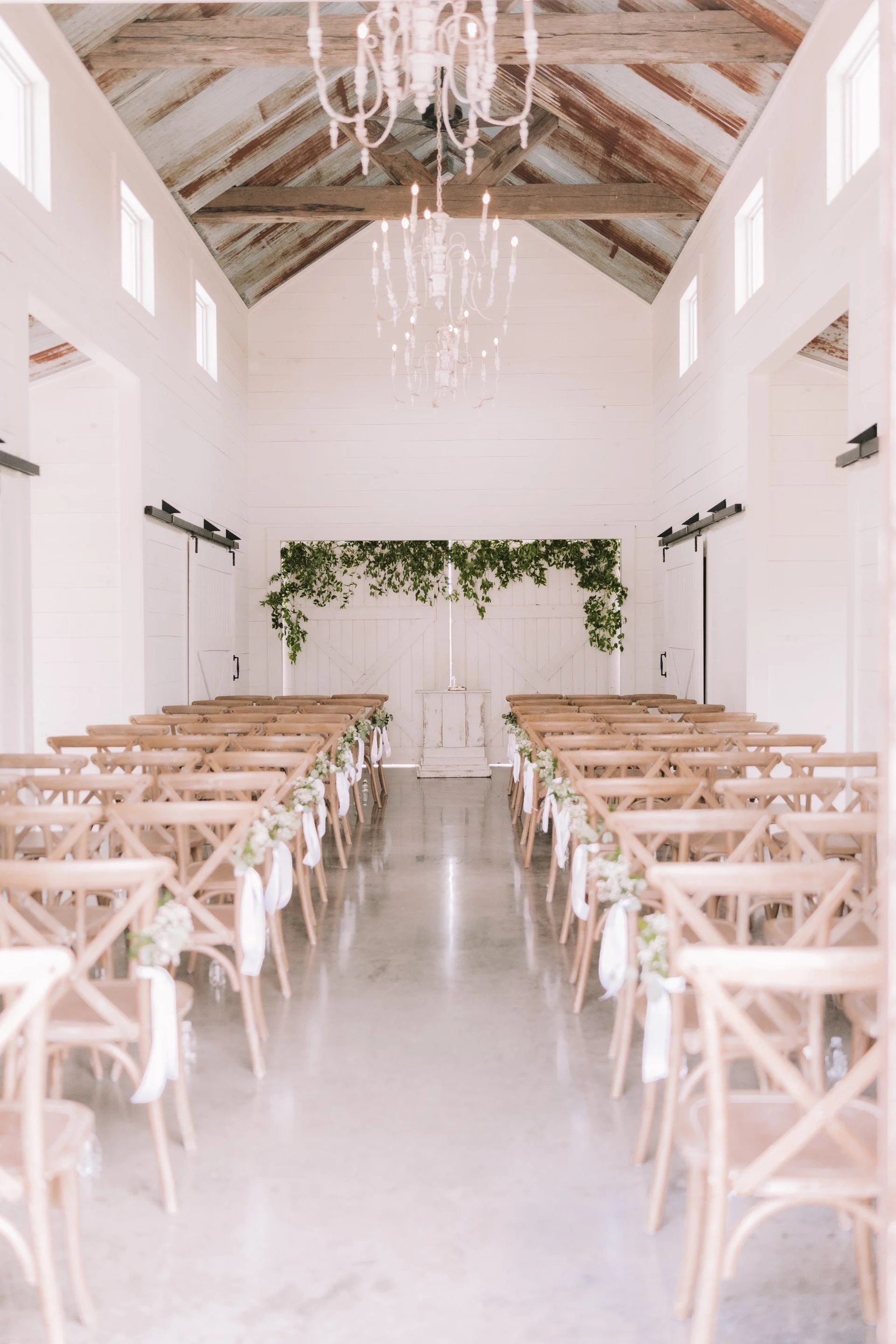Wedding ceremony setup in a rustic, white barn with rows of wooden chairs decorated with white flowers and greenery, a white wooden backdrop, a chandelier hanging from the ceiling, and barn doors at the front.