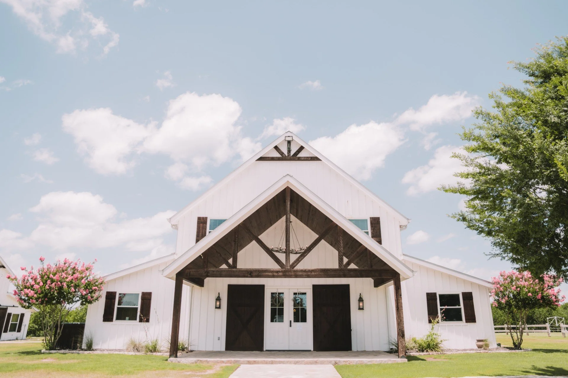 Front view of a white barn-style building with a peaked roof, dark wooden accents, and an open entryway, surrounded by green grass, trees, and pink flowering bushes, under a partly cloudy sky.