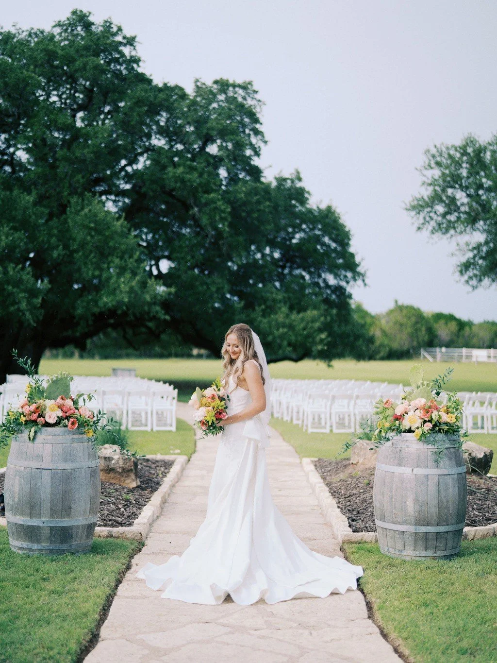 A bride in a white wedding dress holding a bouquet, standing on a stone pathway outdoors, lined with large wooden barrels filled with pink and peach flowers, with white chairs arranged in rows in the background, under a large tree.