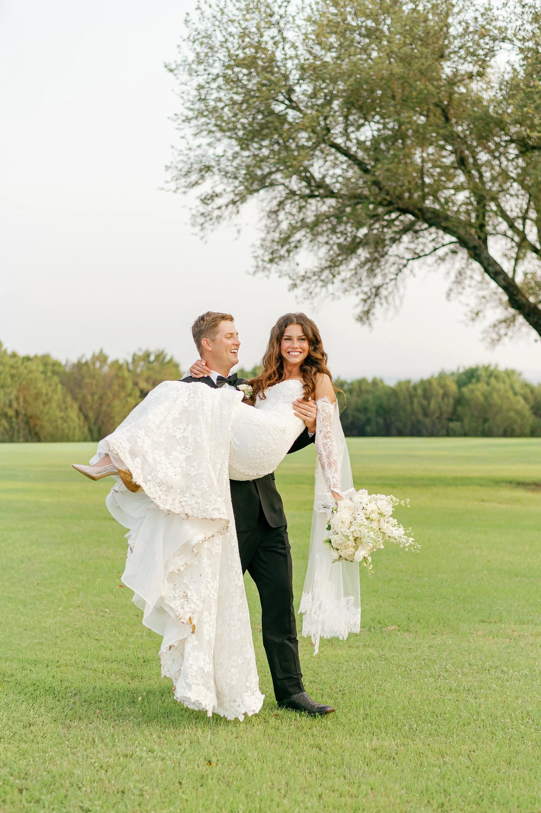 A newlywed couple outdoors on a grassy field, with the groom carrying the bride. The bride is holding a bouquet of white flowers, and both are smiling with trees in the background.