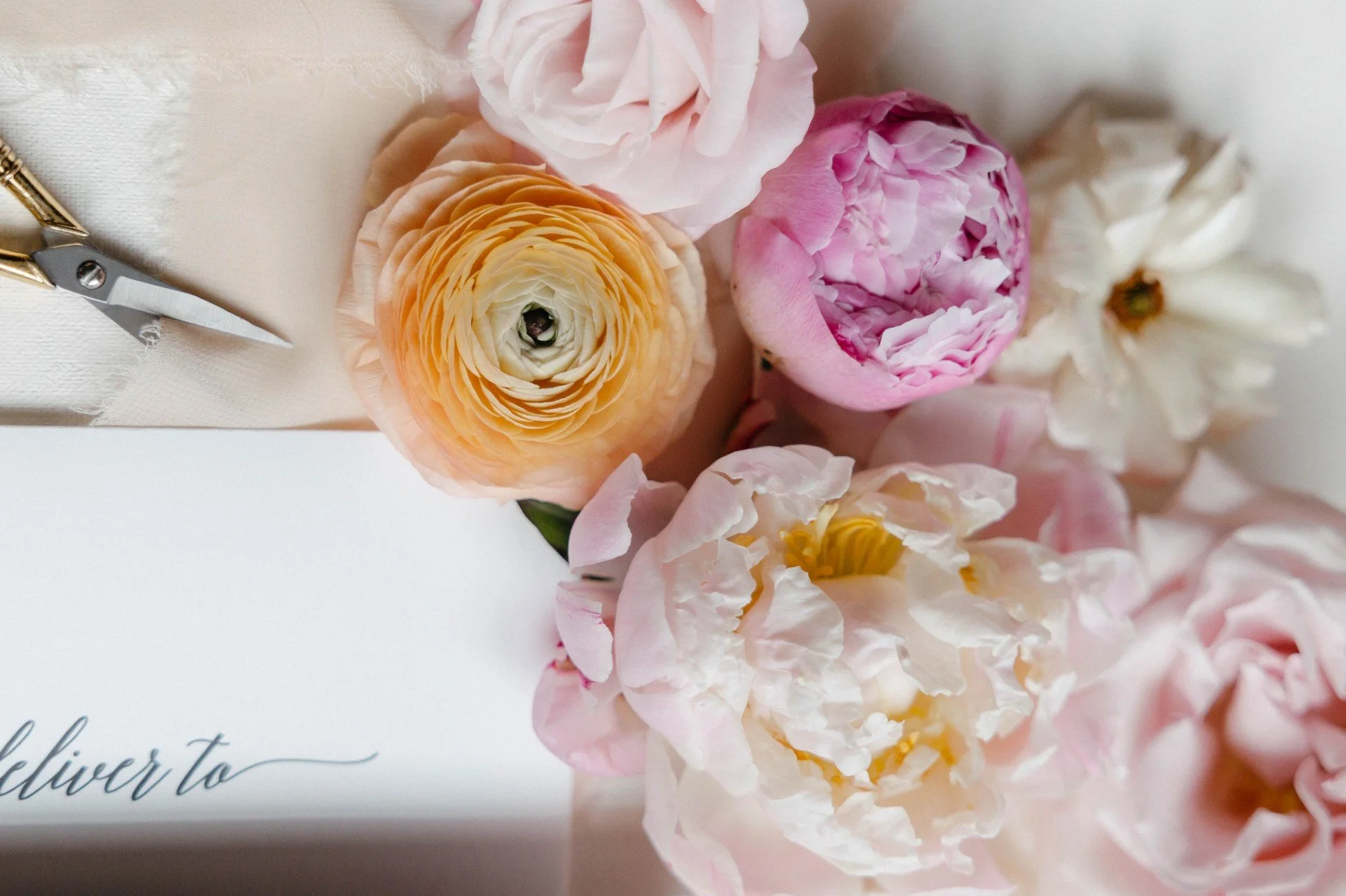 Close-up of pink and yellow ranunculus and peony flowers on a white background with a partially visible card with the word 'deliver to' written in black cursive.