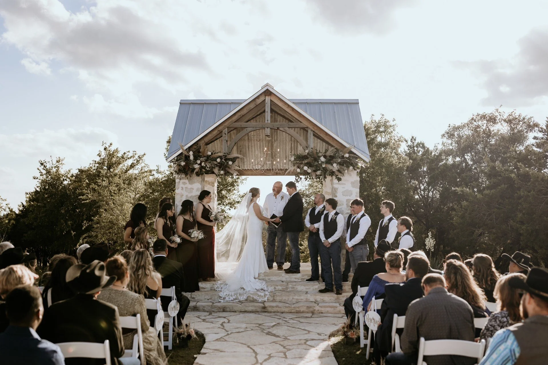 Wedding ceremony taking place outdoors under a wooden chapel with a metal roof, decorated with floral arrangements, in front of a large crowd of guests.