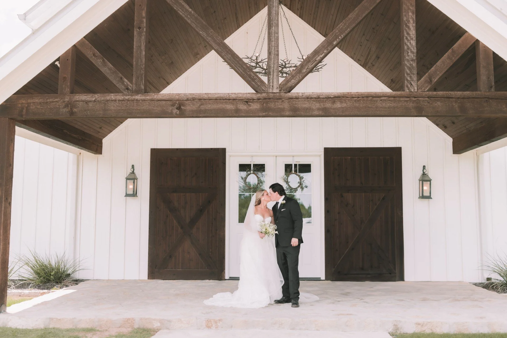 Bride and groom sharing a kiss under a rustic barn archway during their wedding, with white walls, dark wooden doors, and decorative wreaths in the background.