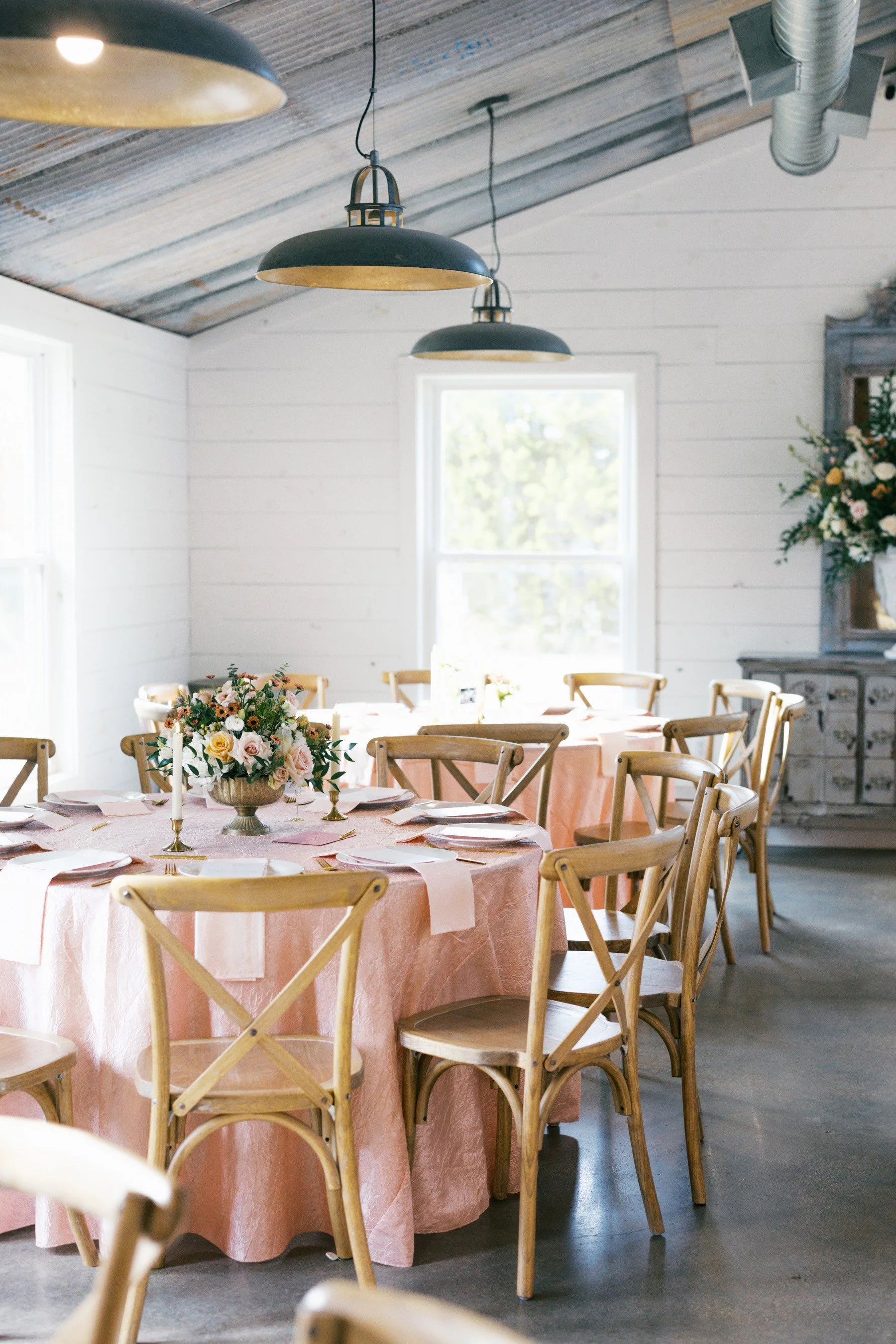 A dining room with a round table covered in a pink tablecloth. The table is set with white plates, gold utensils, and a large floral centerpiece. Wooden chairs surround the table. The room has white paneled walls, large windows, and industrial-style pendant lights hanging from a wooden ceiling with exposed ductwork.