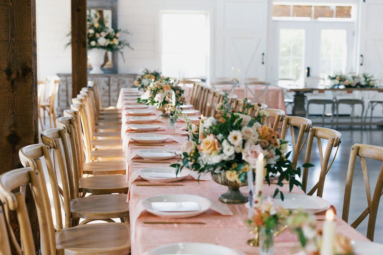 A long rectangular dining table set for a celebration in a bright room with large windows. The table has pink tablecloths, white plates, floral centerpieces, and candles. Wooden chairs surround the table, and additional floral arrangements are visible in the background.