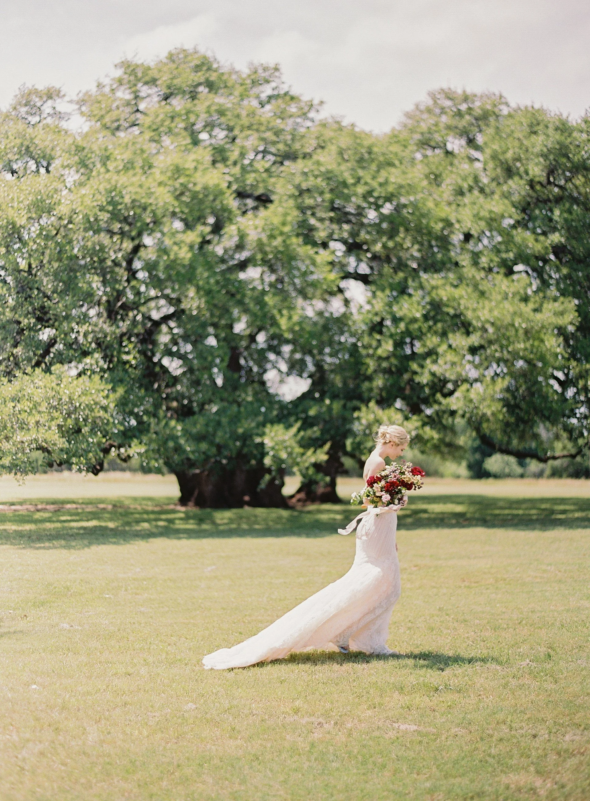 A woman in a wedding dress holding a bouquet of flowers, outdoors under a large leafy tree on a sunny day.