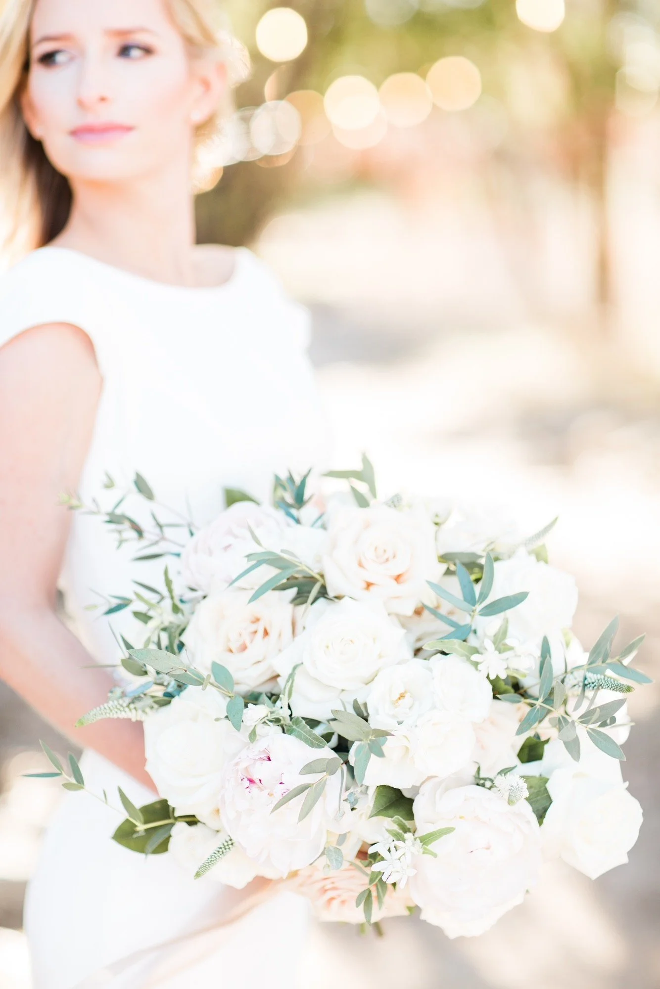 A woman holding a large bouquet of white and blush roses with green foliage at an outdoor event during daytime.
