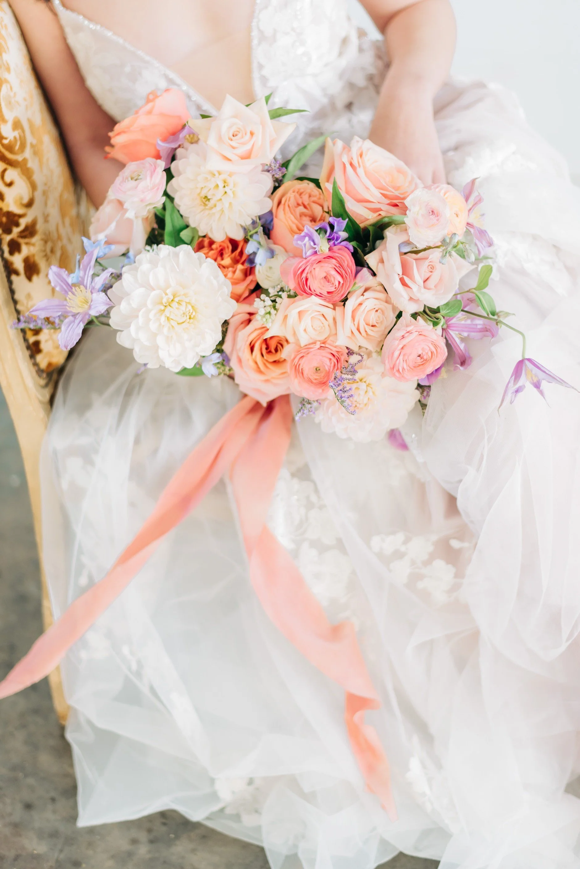 Close-up of a bride holding a bouquet of pastel-colored flowers, including roses and other blooms, with a peach-colored ribbon, resting on a surface with sheer fabric and lace details.