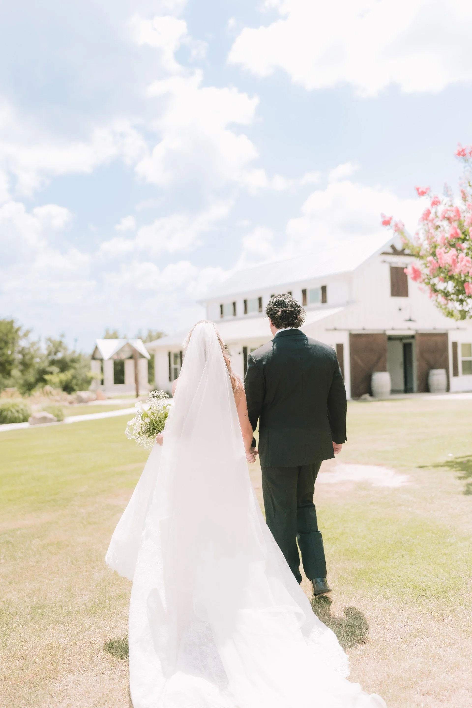 A bride and groom walking hand in hand outdoors on a sunny day with a large white house, trees, and a pink flowering tree in the background.