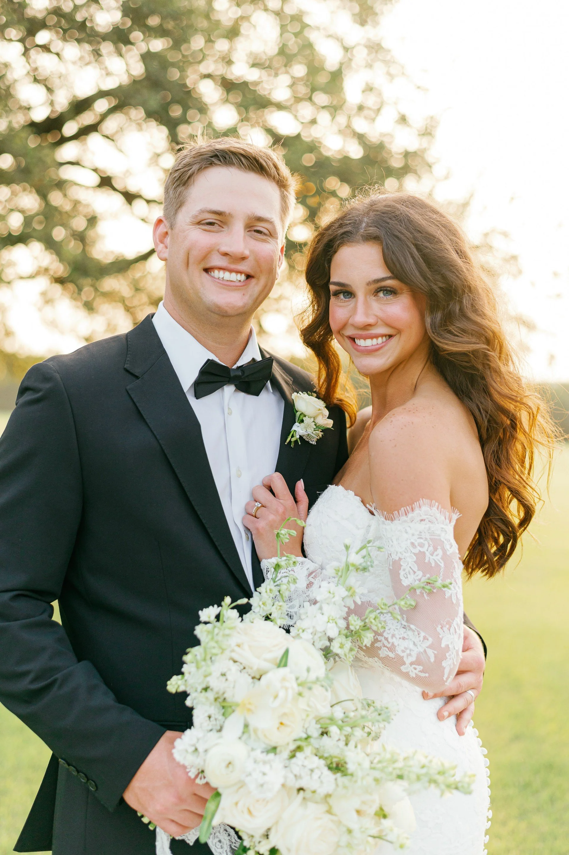 A smiling couple on their wedding day standing outdoors, with trees in the background and sunlight filtering through the leaves, the groom in a black tuxedo and the bride in a white lace wedding dress holding a bouquet of white flowers.