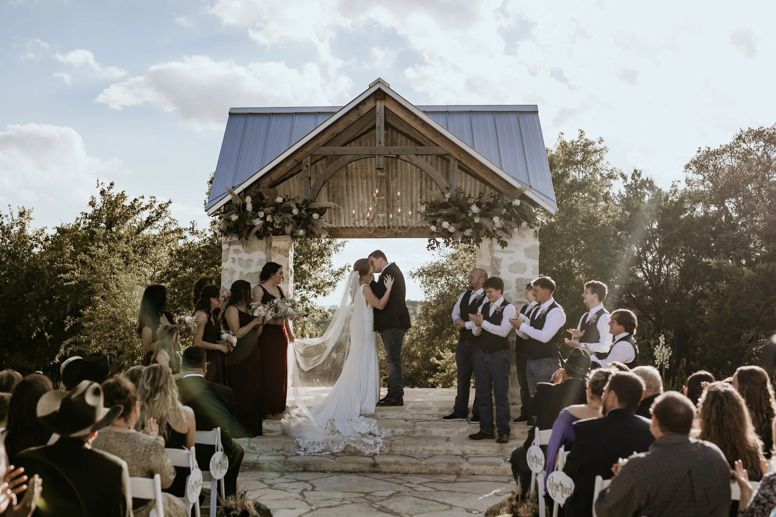 A wedding ceremony taking place outdoors under a rustic stone and wood pavilion with a metal roof, decorated with flowers. The bride and groom are kissing, with the officiant and bridesmaids and groomsmen standing nearby. Guests are seated and watching attentively.