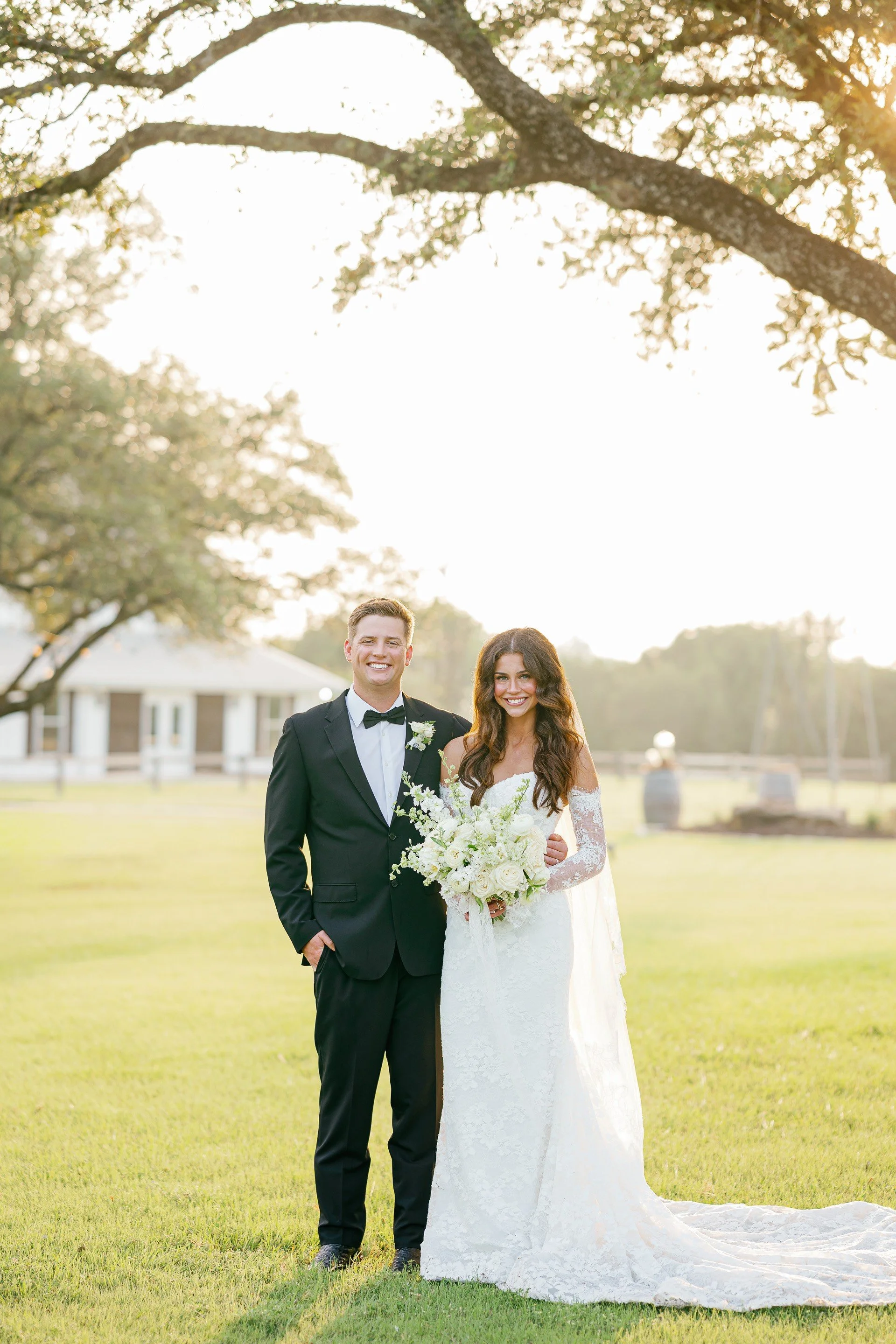 A newlywed couple standing on a green lawn during sunset, with trees and a house in the background. The groom is in a black tuxedo with a bow tie, and the bride is in a white lace wedding dress holding a bouquet of white flowers. Both are smiling.