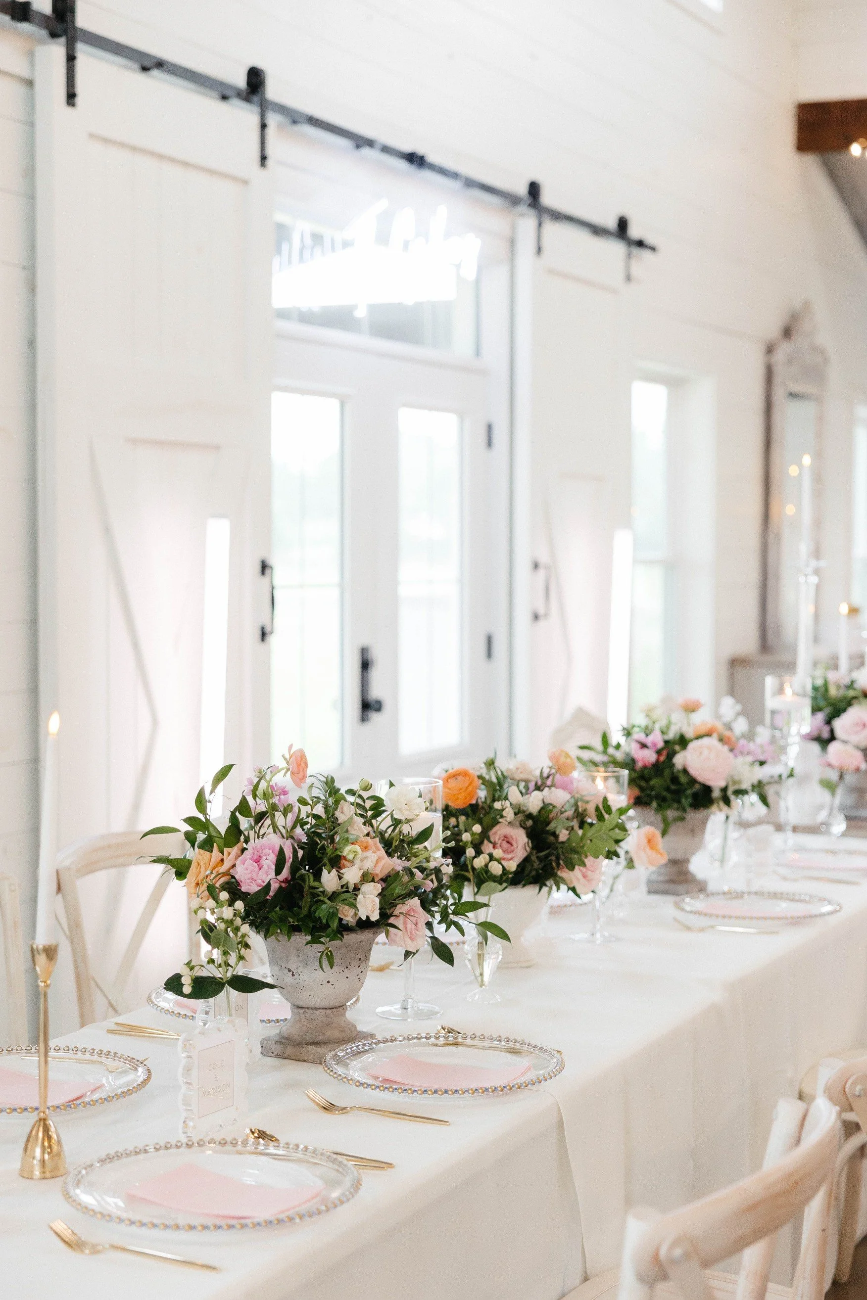 A decorated table with floral centerpieces, pink and white flower arrangements in vases, and elegant tableware, set for a celebration or wedding in a bright, airy room with large windows and white walls.