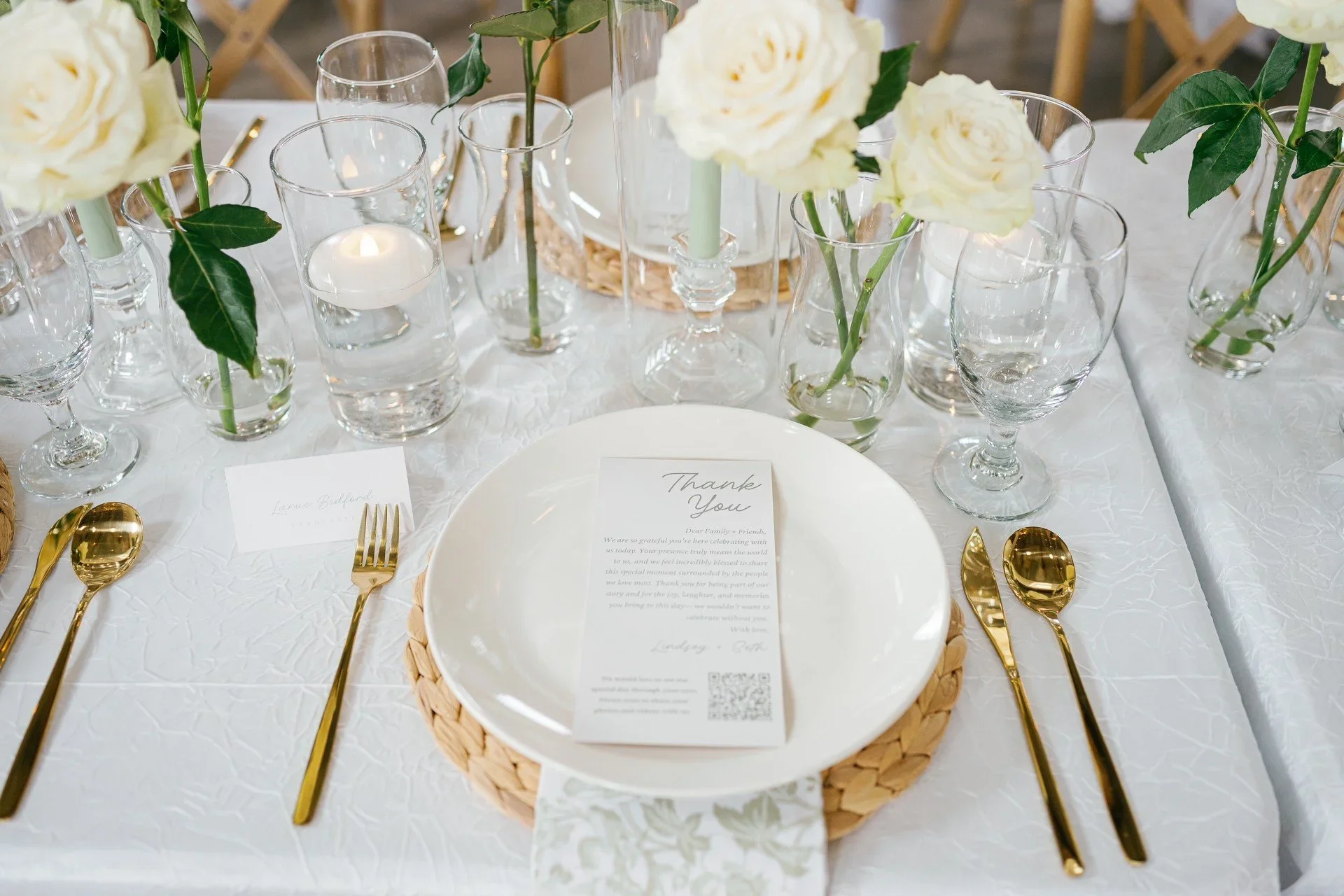 A table setting for a special occasion with white roses in glass vases, gold cutlery, and a white textured tablecloth. There are clear glasses containing candles and a white plate with a printed thank you note.