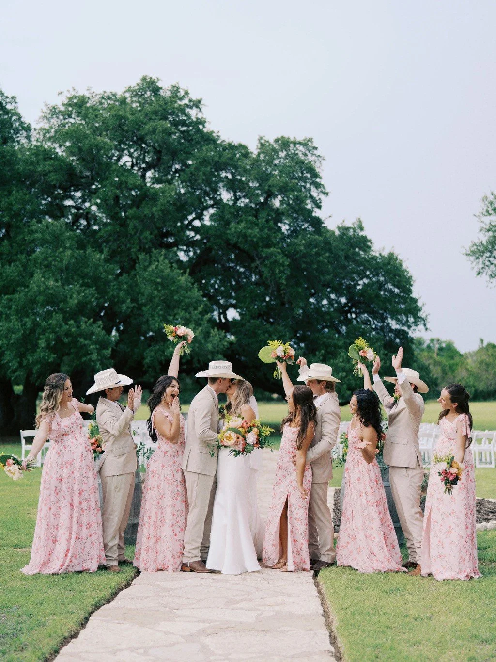 A wedding party outdoors on a grassy area, with bridesmaids in pink floral dresses and groomsmen in beige suits and hats, celebrating and raising bouquets in the air.