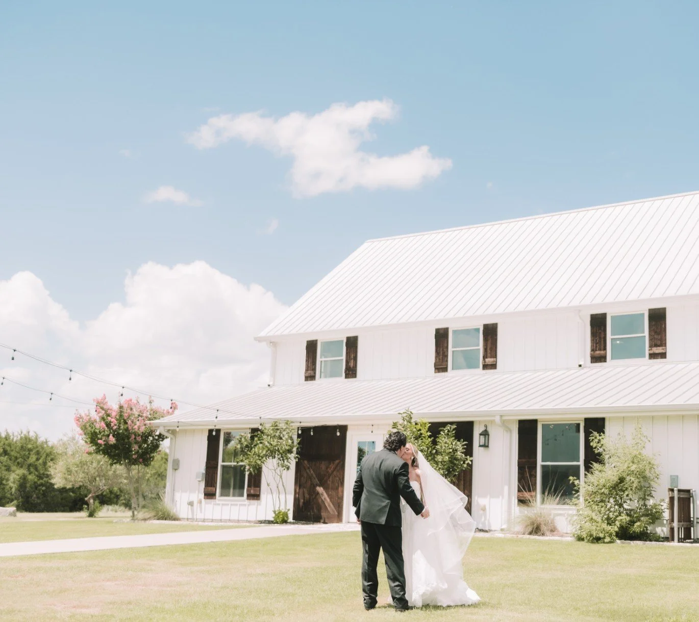A bride and groom sharing a kiss outside a white barn with string lights and trees, under a partly cloudy sky.