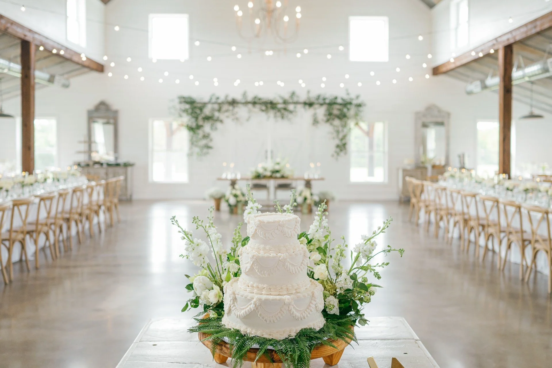 Wedding cake with white frosting and floral decorations in front of a decorated reception hall with string lights and seating arrangements.