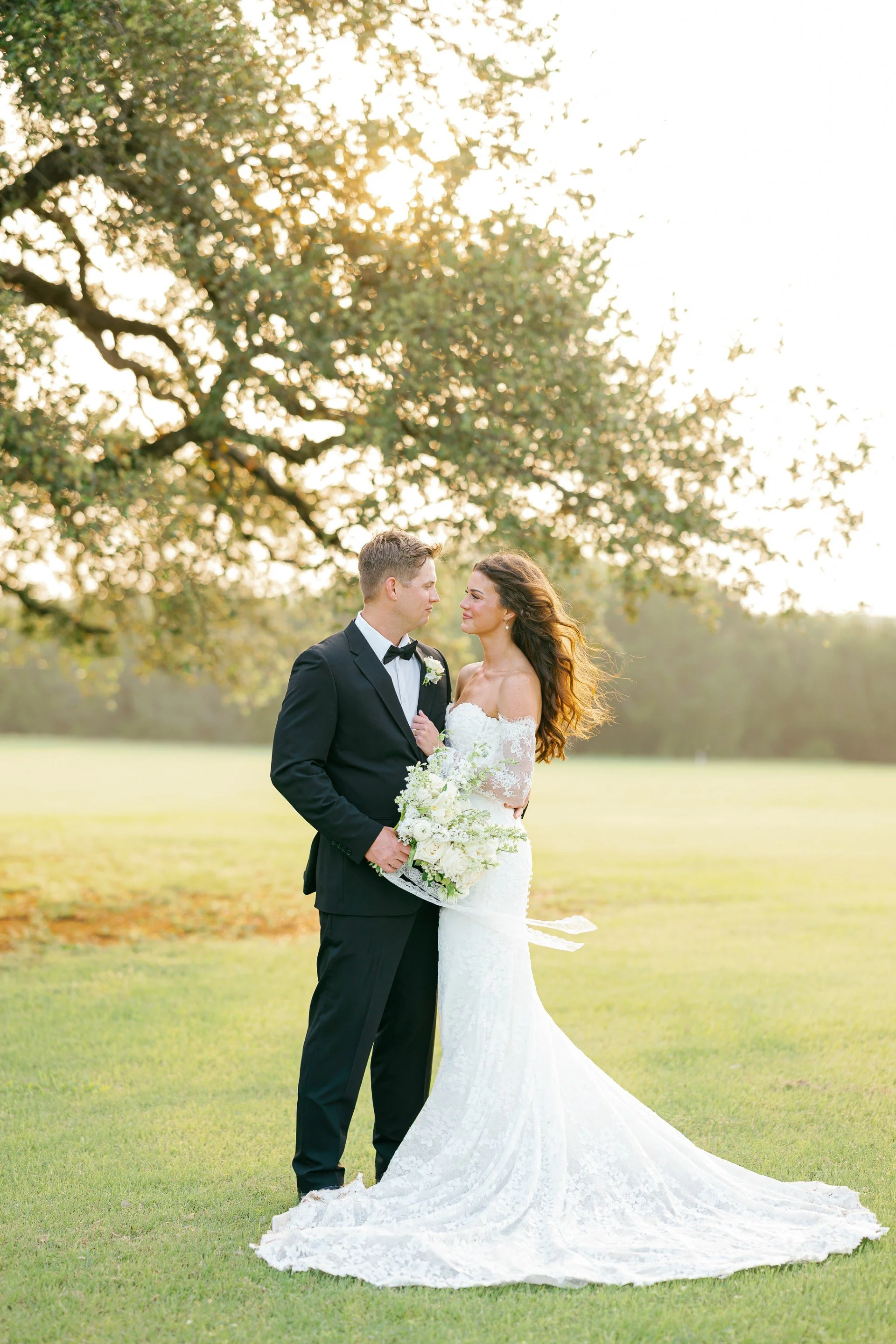 A bride and groom stand face to face outdoors, with the groom in a black tuxedo and the bride in a strapless white wedding gown holding a bouquet of white flowers, under a large tree with sunlight filtering through the leaves, during sunset.