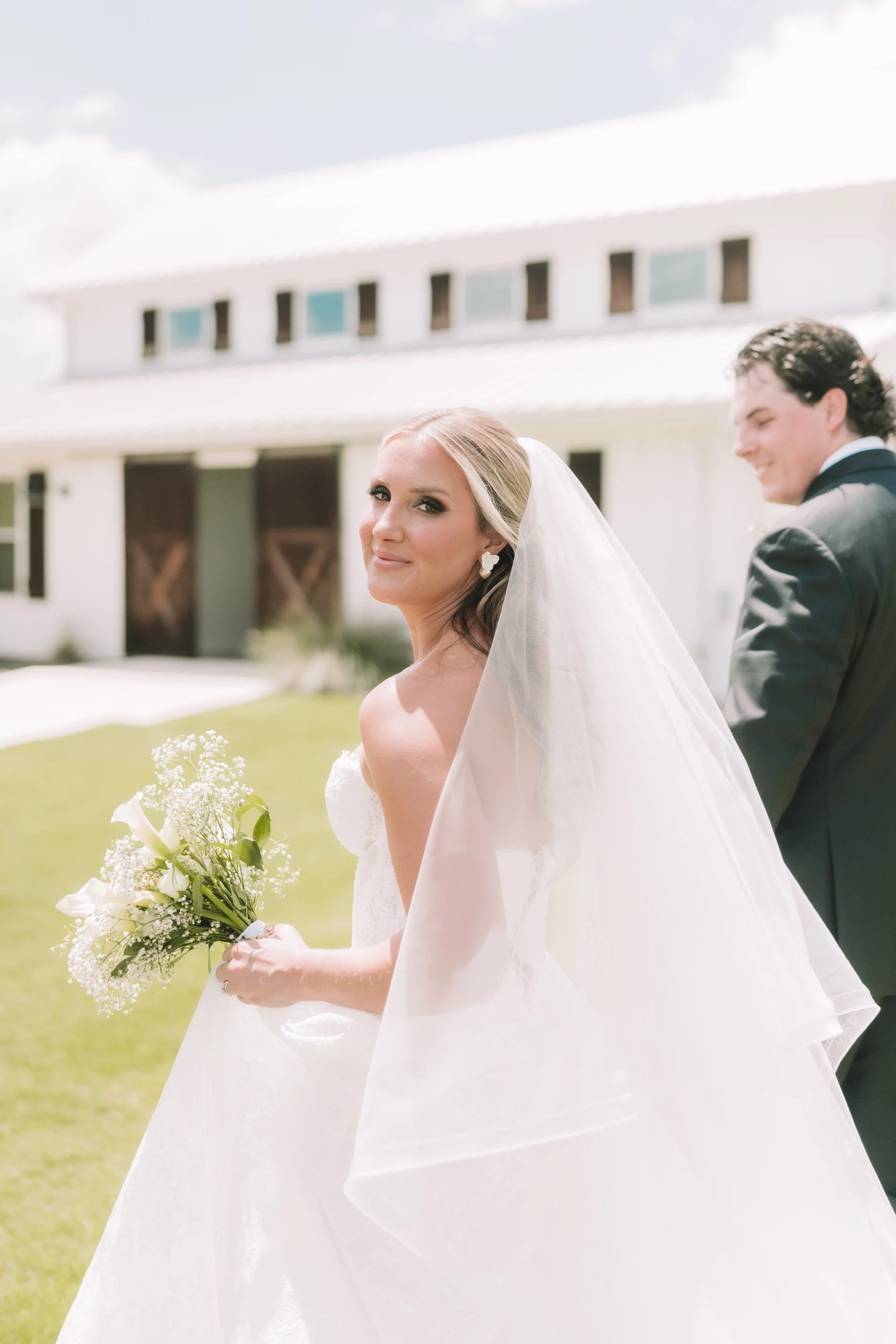 A bride in a white wedding gown holding a bouquet of white flowers, standing outdoors near a white barn or house with a metal roof, smiling at the camera. Groom stands beside her, slightly blurred, wearing a black tuxedo. The scene is bright with a partly cloudy sky.
