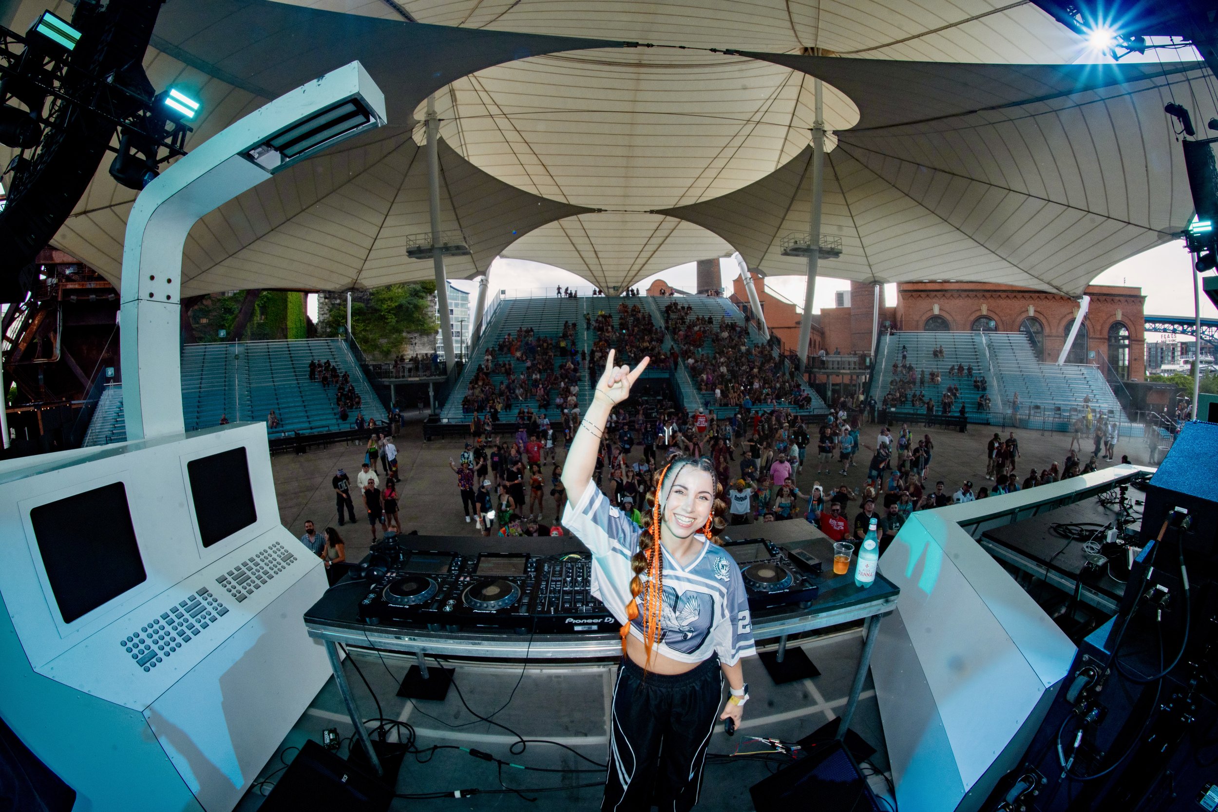Female DJ smiling and making a peace sign with her hand at an outdoor music event, with a large crowd and stage in the background.