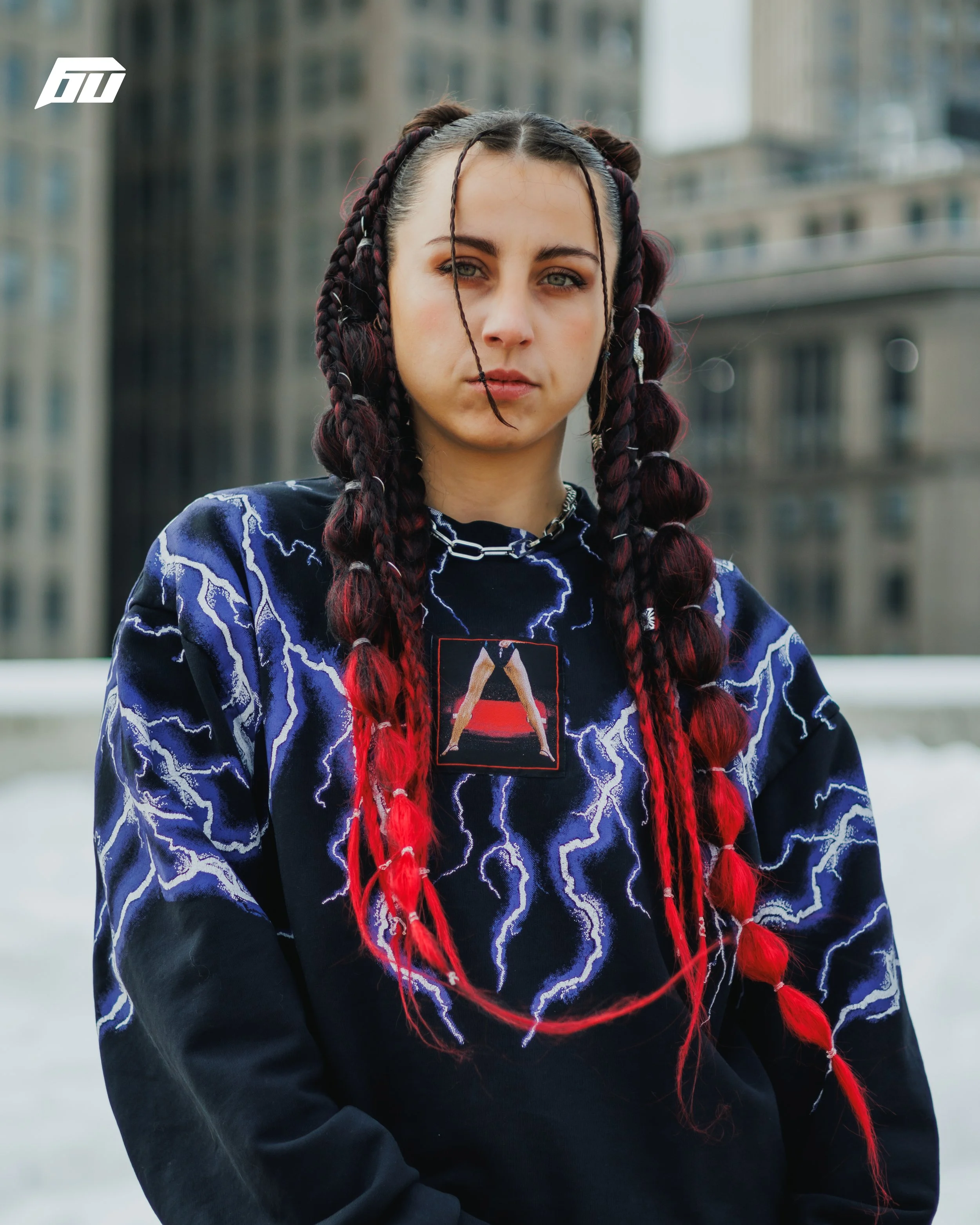A young woman with braided hair, wearing a black sweatshirt with blue lightning patterns and a chain necklace, stands outdoors in front of blurred city buildings.