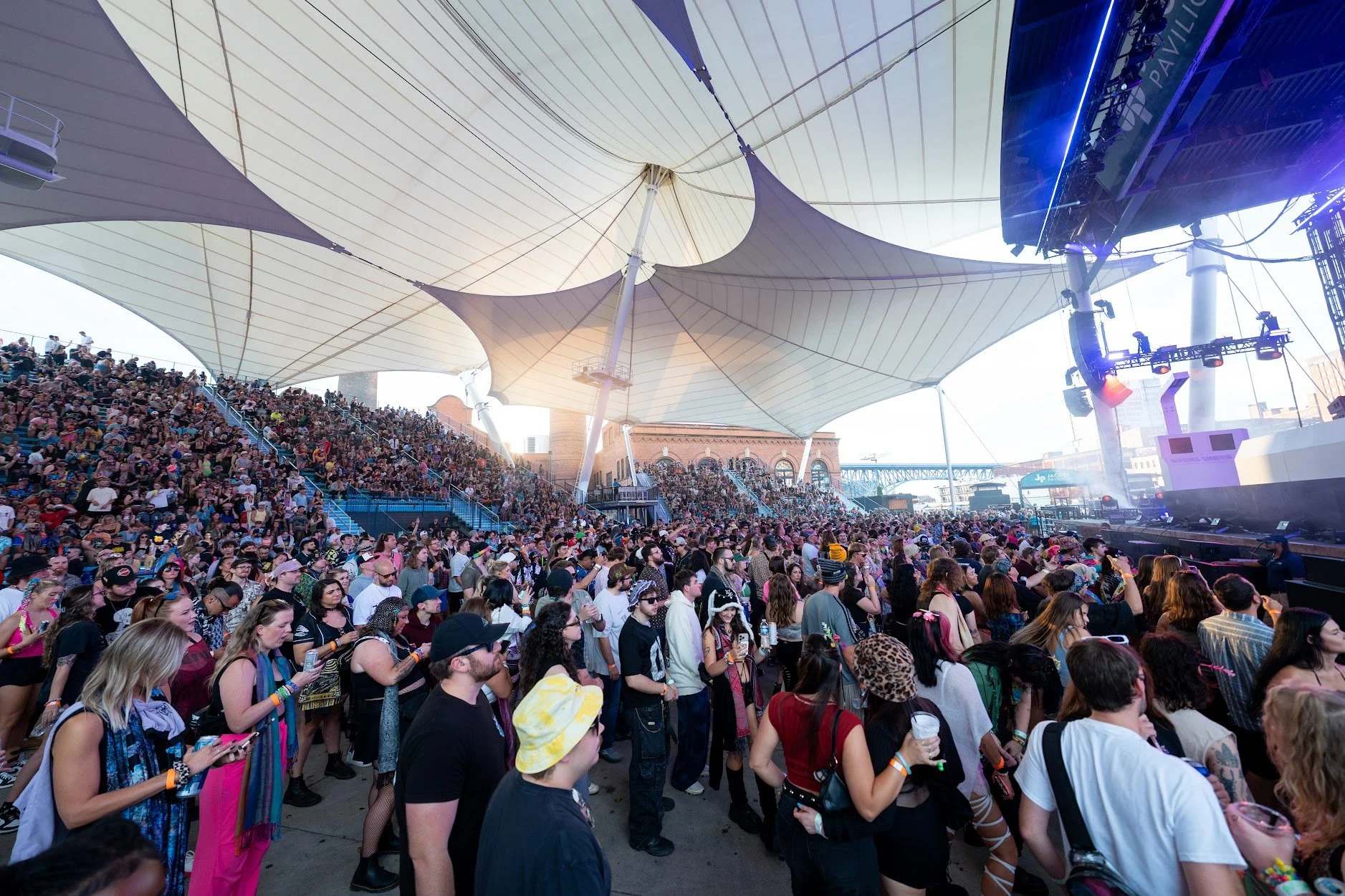 Large crowd at an outdoor music concert under a tent with white fabric sails, stage with speakers and lighting on the right, and people enjoying the event.