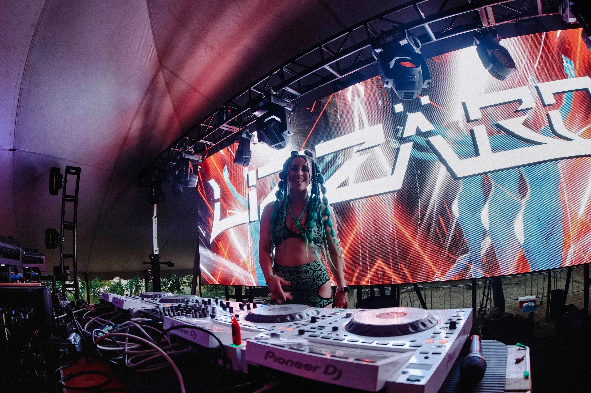 A person standing behind DJ equipment at an outdoor electronic music event, with a large LED screen displaying vibrant graphics in the background.