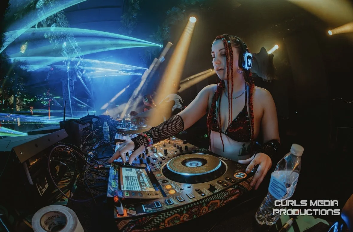 A female DJ with red braided hair wearing headphones, a black crop top, and fishnet gloves, is mixing music at a club with colorful lighting and laser effects.