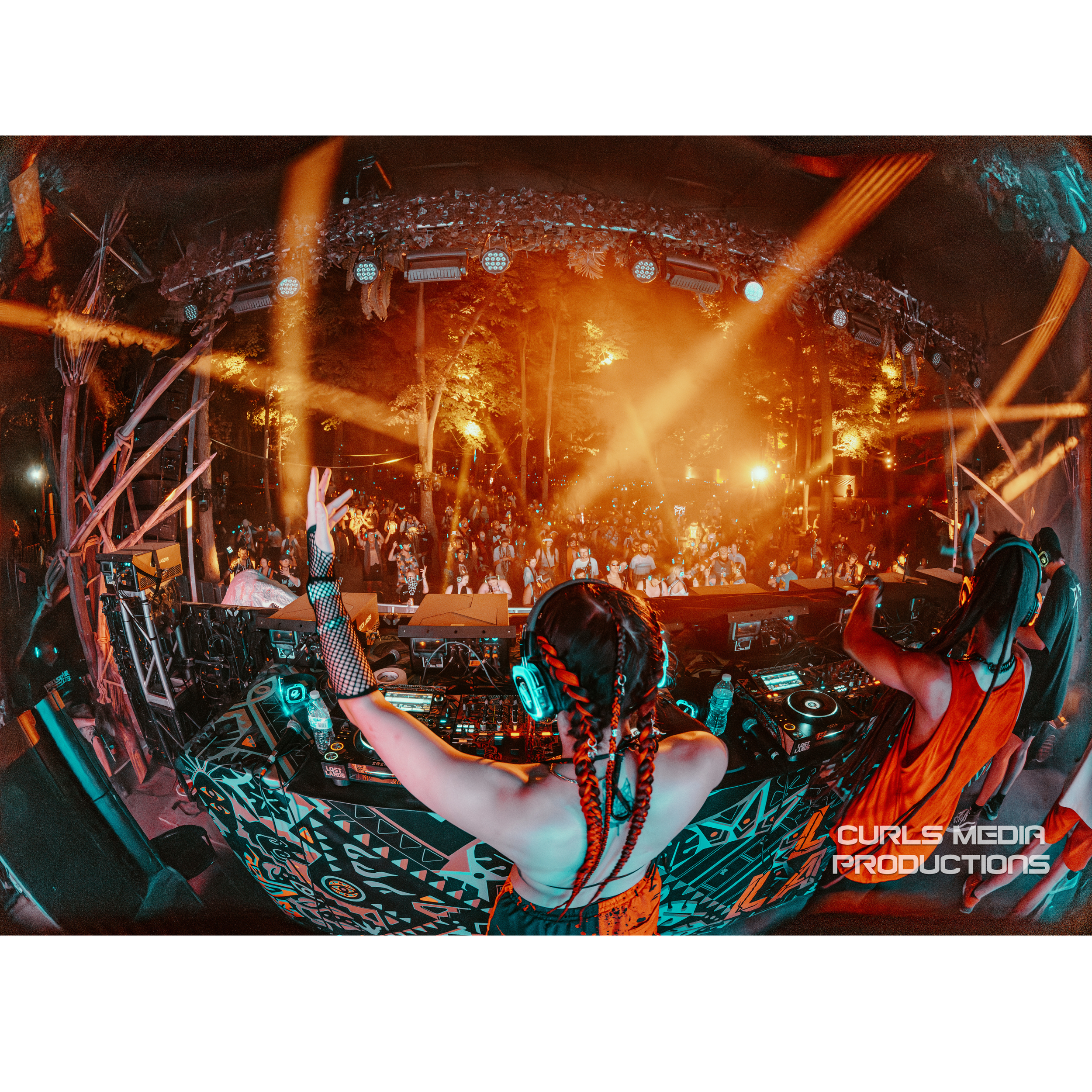 A festival scene showing a crowd of people under a decorated outdoor stage with bright orange lights and trees in the background. A DJ woman with braids and headphones is at the mixing station.