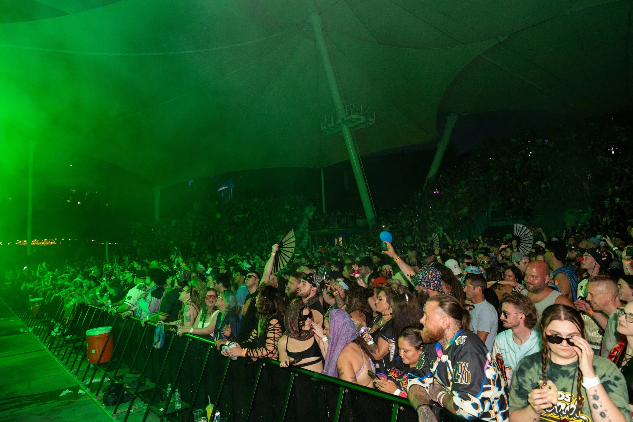 Crowd of people attending a concert or event with some holding fans and wearing colorful clothing, in an indoor venue with a green light on the large ceiling structure.