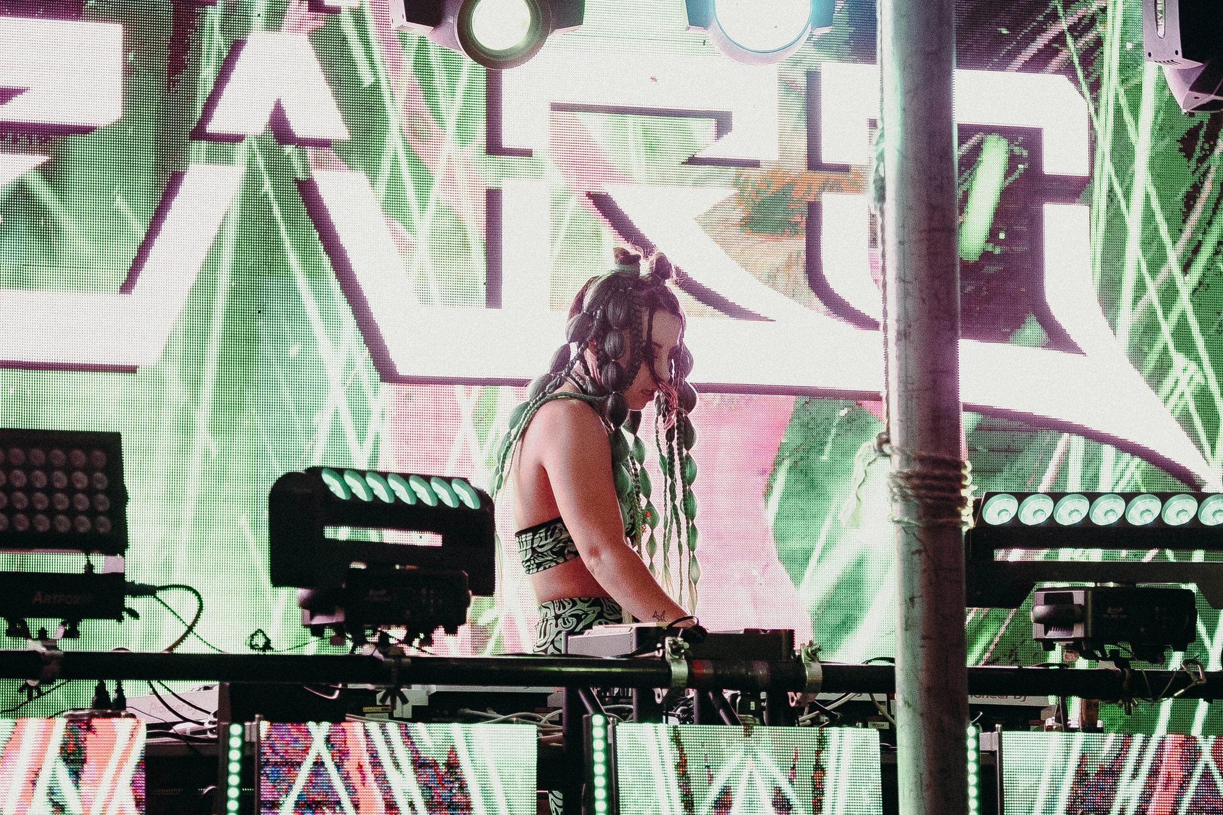 A DJ with long, braided hair performing at a music event, with a vibrant, colorful, neon-lit backdrop displaying geometric and abstract patterns.