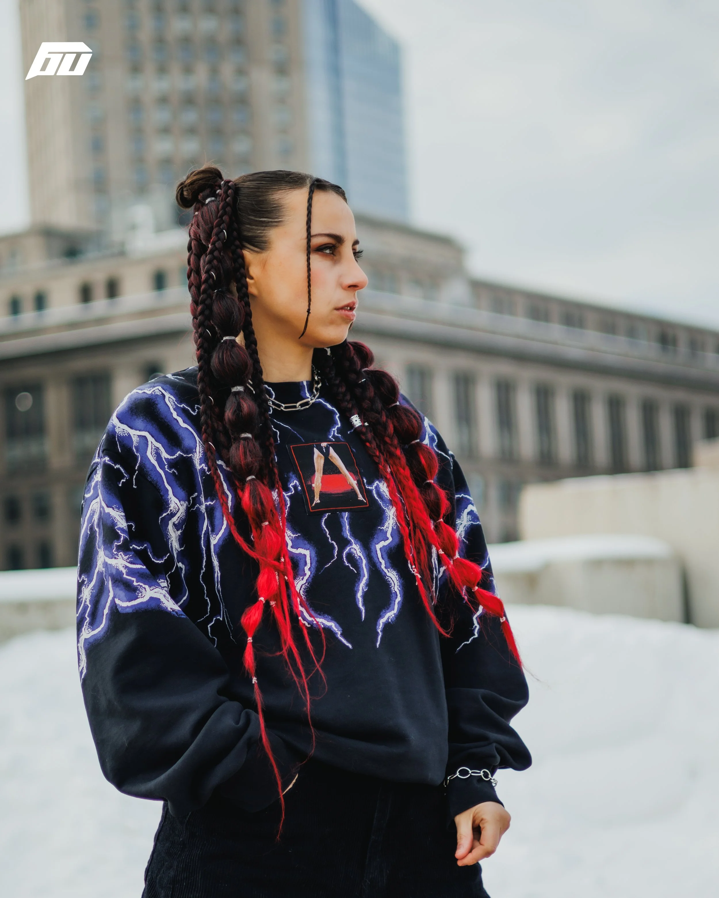 A woman with long, braided hair colored with red and black, wearing a blue and black storm pattern sweatshirt and black pants, standing outdoors in an urban area with tall buildings and snow on the ground.